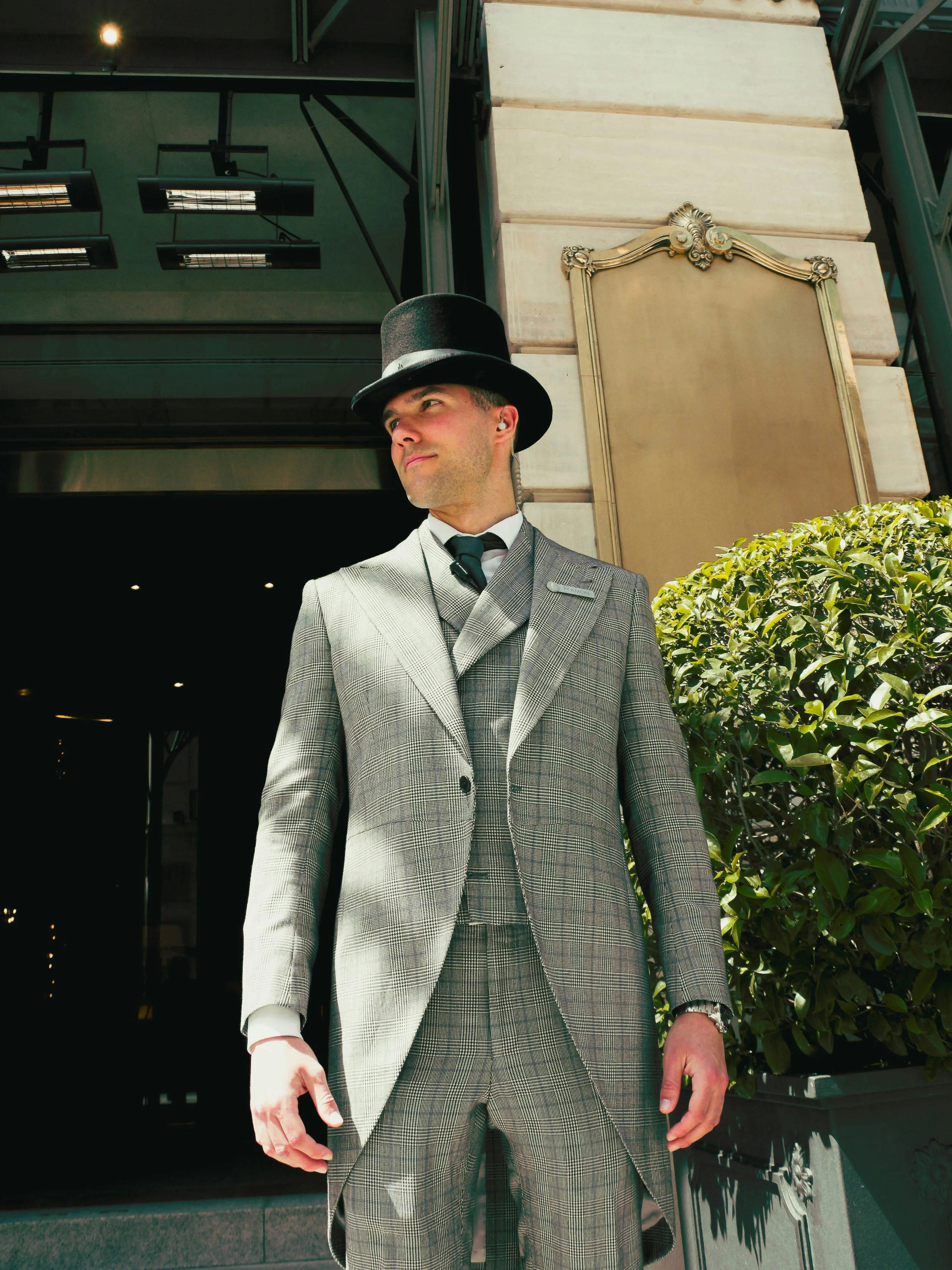 A man in a plaid suit and top hat standing outside near a building with green plants and a gold framed sign.