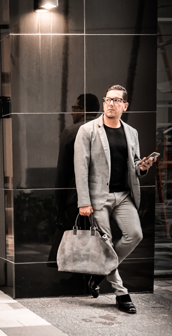 Man in gray suit holding a gray bag and a smartphone, standing against a black tiled wall, reflecting his image.