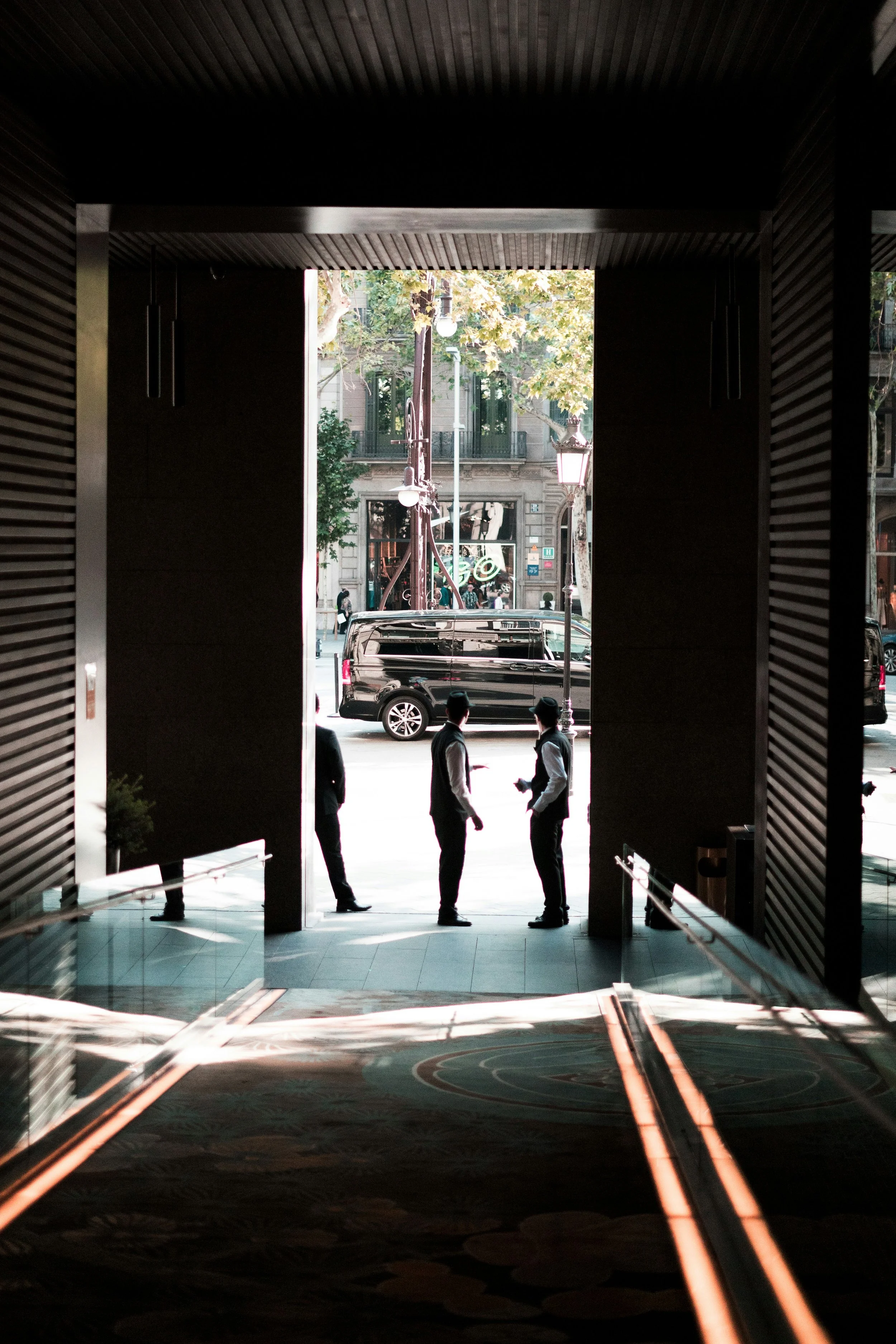 People walking outdoors through an archway, city scene with vehicles, trees, and buildings visible in the background.