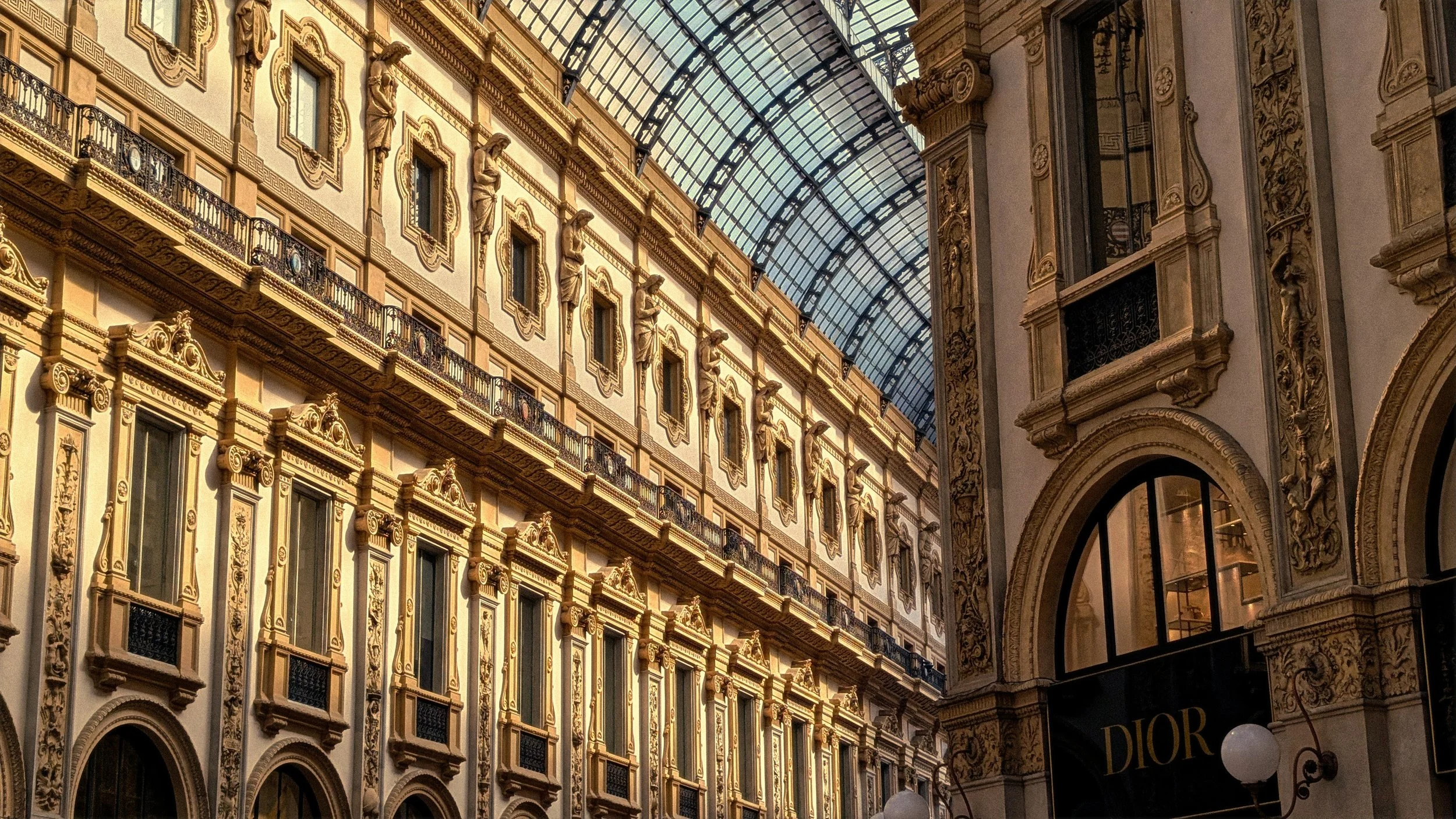 Interior view of a luxurious shopping arcade with ornate gold and cream facades, decorative statues, and a curved glass ceiling.