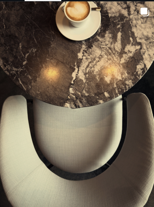 A marble-topped round table with a cup of coffee on a white saucer, seen from a top-down perspective, with two beige upholstered chairs around the table.