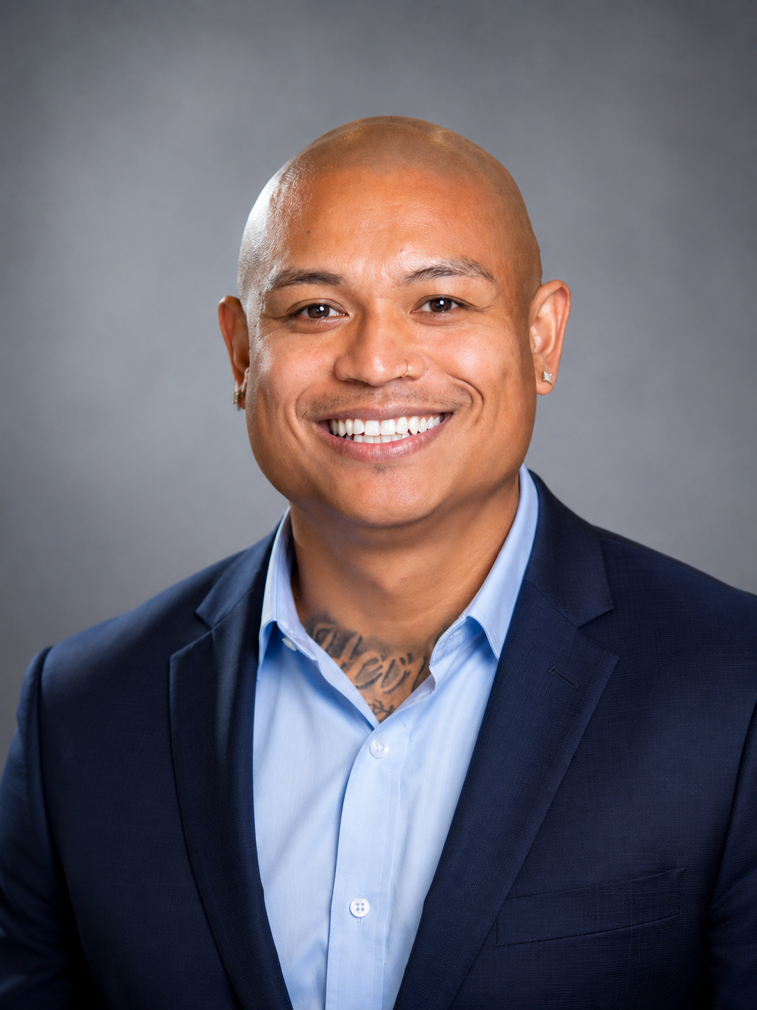 Headshot of a smiling man with a shaved head, wearing a dark suit and light blue shirt, against a gray background.