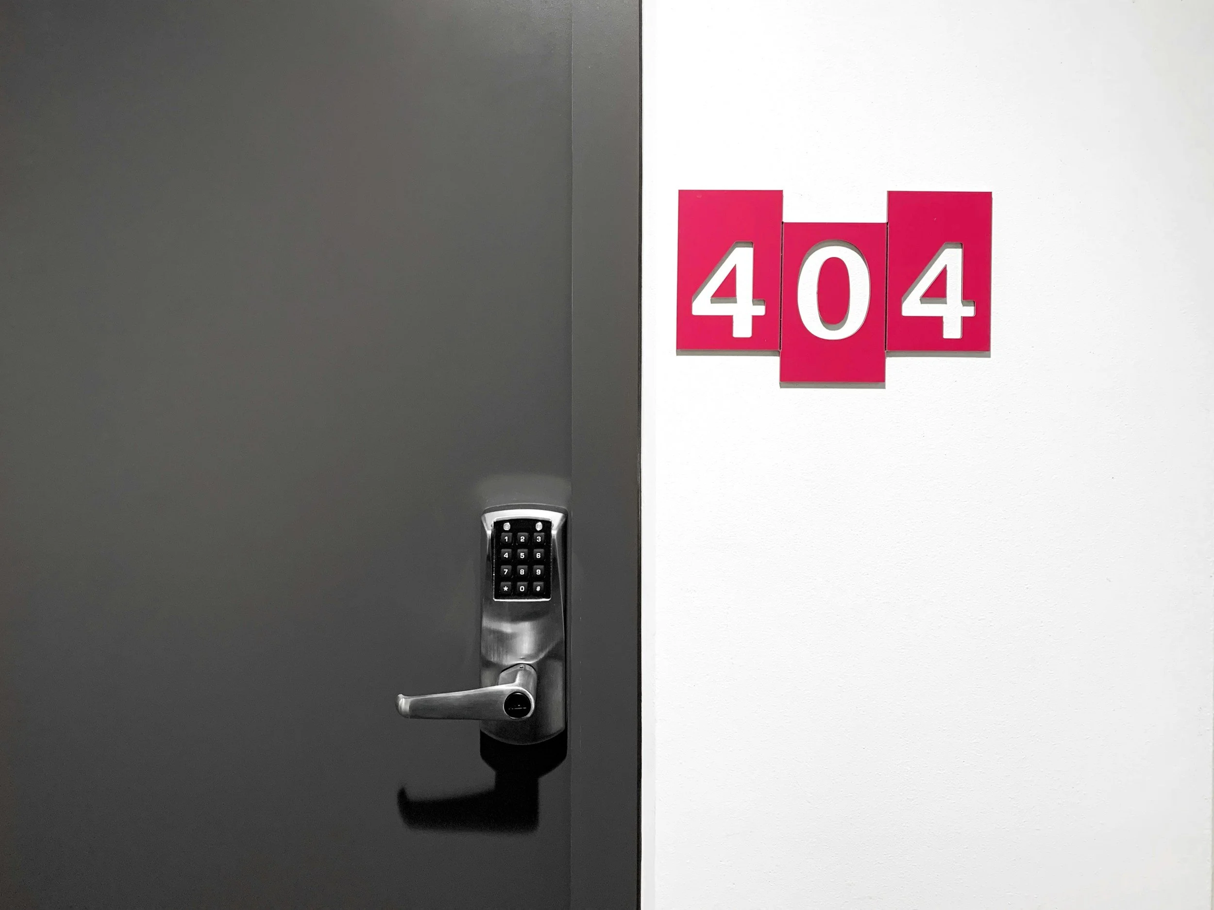 A dark gray door with a metallic handle and keypad lock, next to a white wall with pink and white signage displaying the number 404.