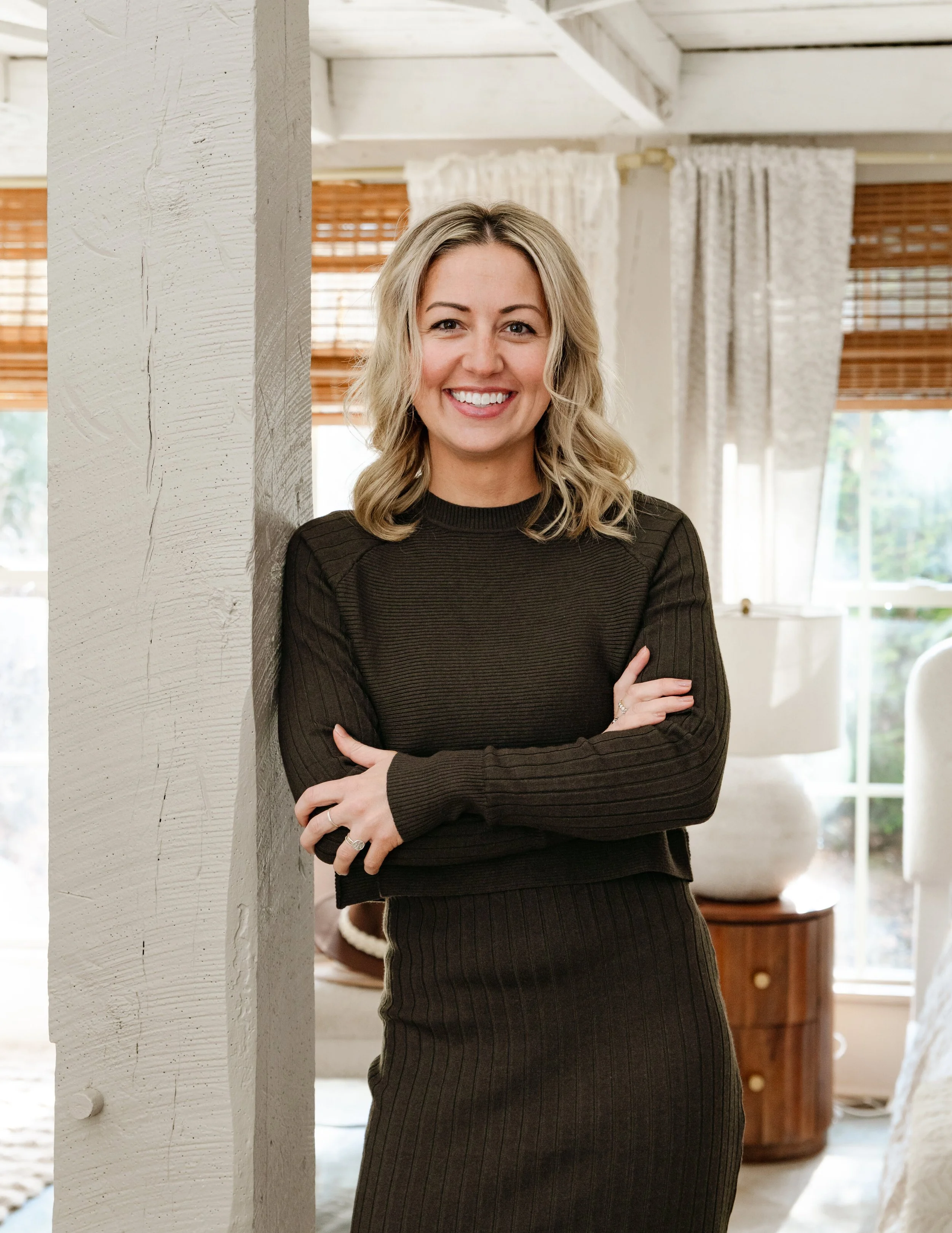 A smiling woman with blonde hair, wearing a black outfit, standing indoors next to a white textured wall with windows, curtains, and wooden blinds in the background.
