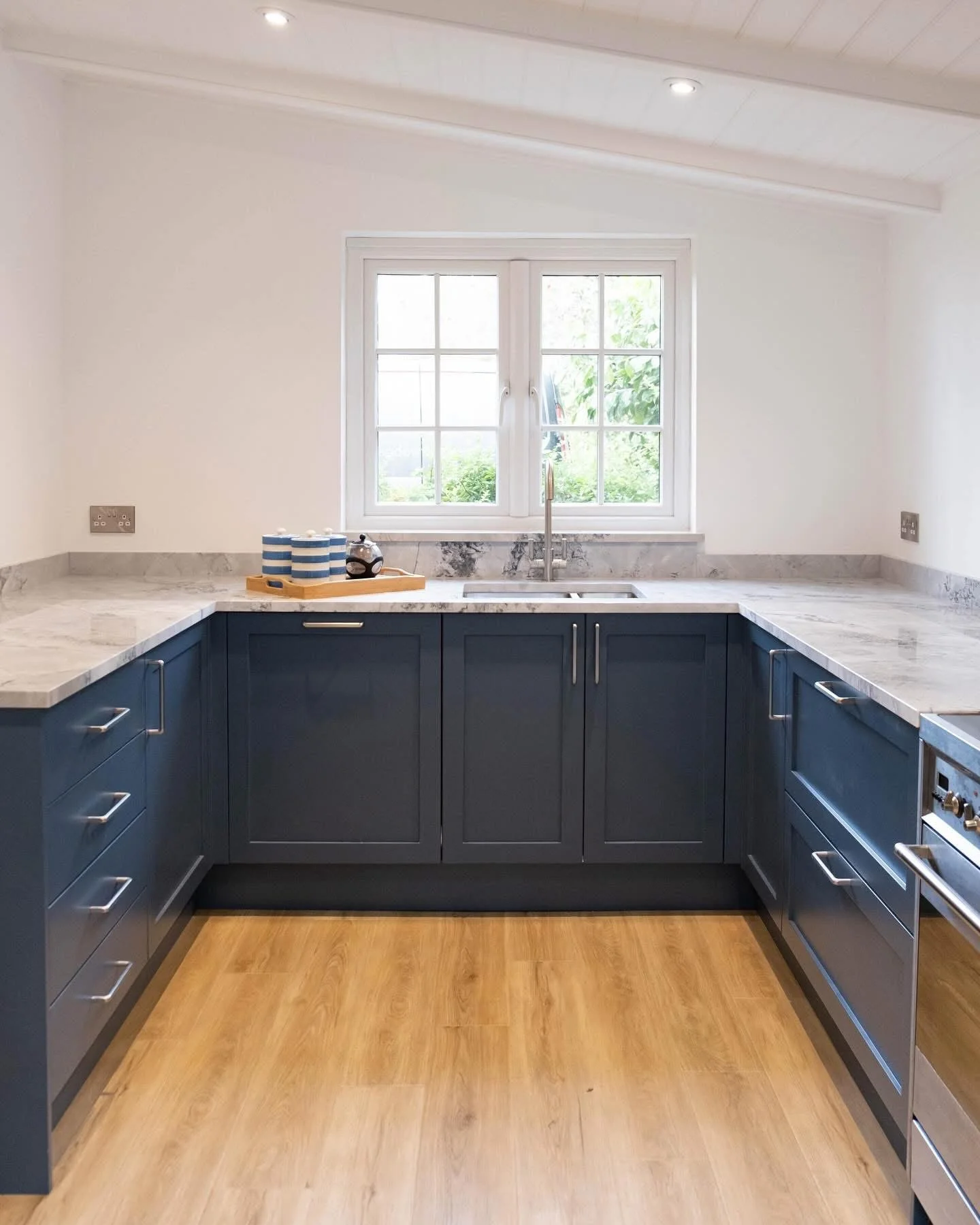 A modern kitchen with blue cabinets, a marble countertop, and a window above the sink, with some dishes and a teapot on a wooden tray.
