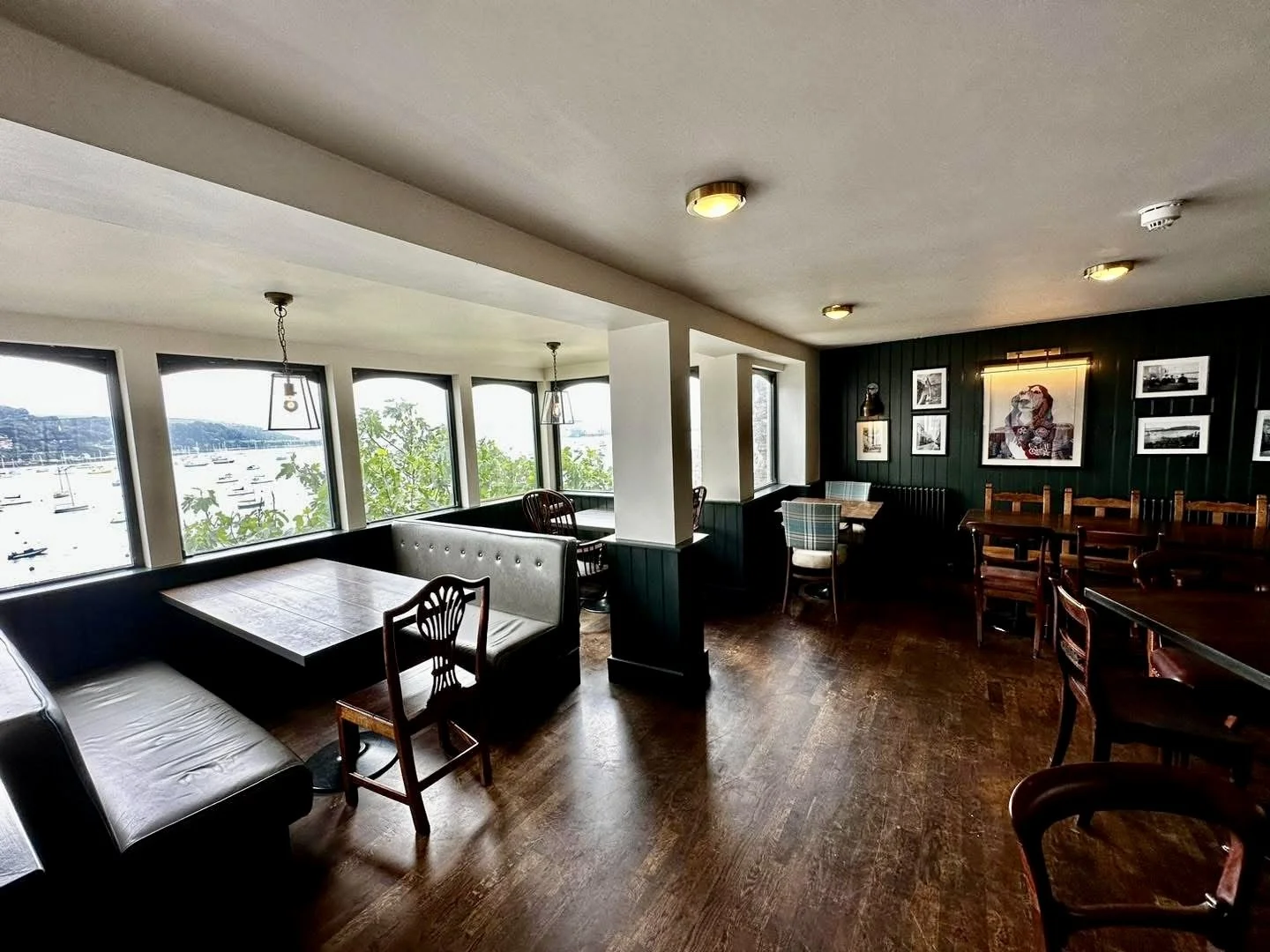 Interior of a restaurant or cafe with wooden tables and chairs, large windows overlooking a marina with boats, and black and white photos on a dark paneled wall.