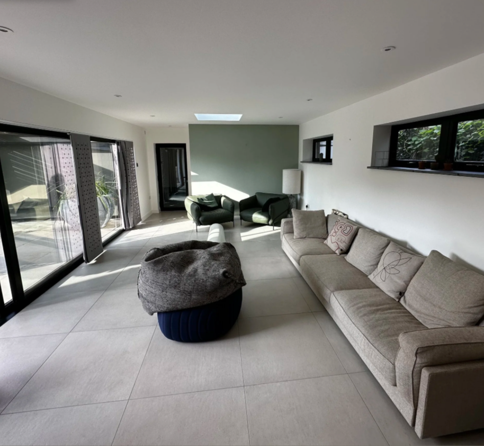 Modern living room with a beige sectional sofa, two green armchairs, sliding glass doors, a ceiling skylight, and small windows with greenery outside.