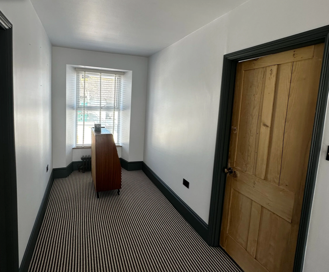 A small, narrow room with white walls, a window with blinds, a wooden dresser, and a wooden door.