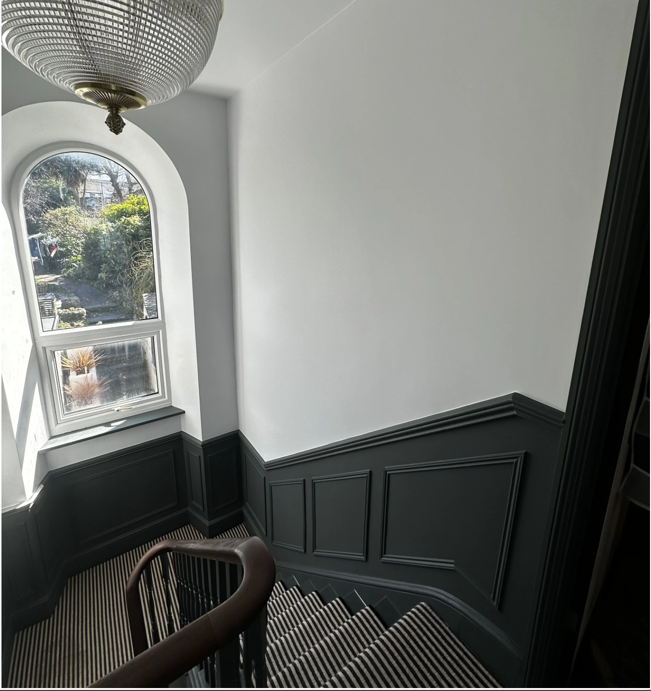 Inside a stairwell landing with a window showing a sunny outdoor garden, dark green wainscoting on the walls, a striped carpeted staircase, and a decorative hanging light fixture.