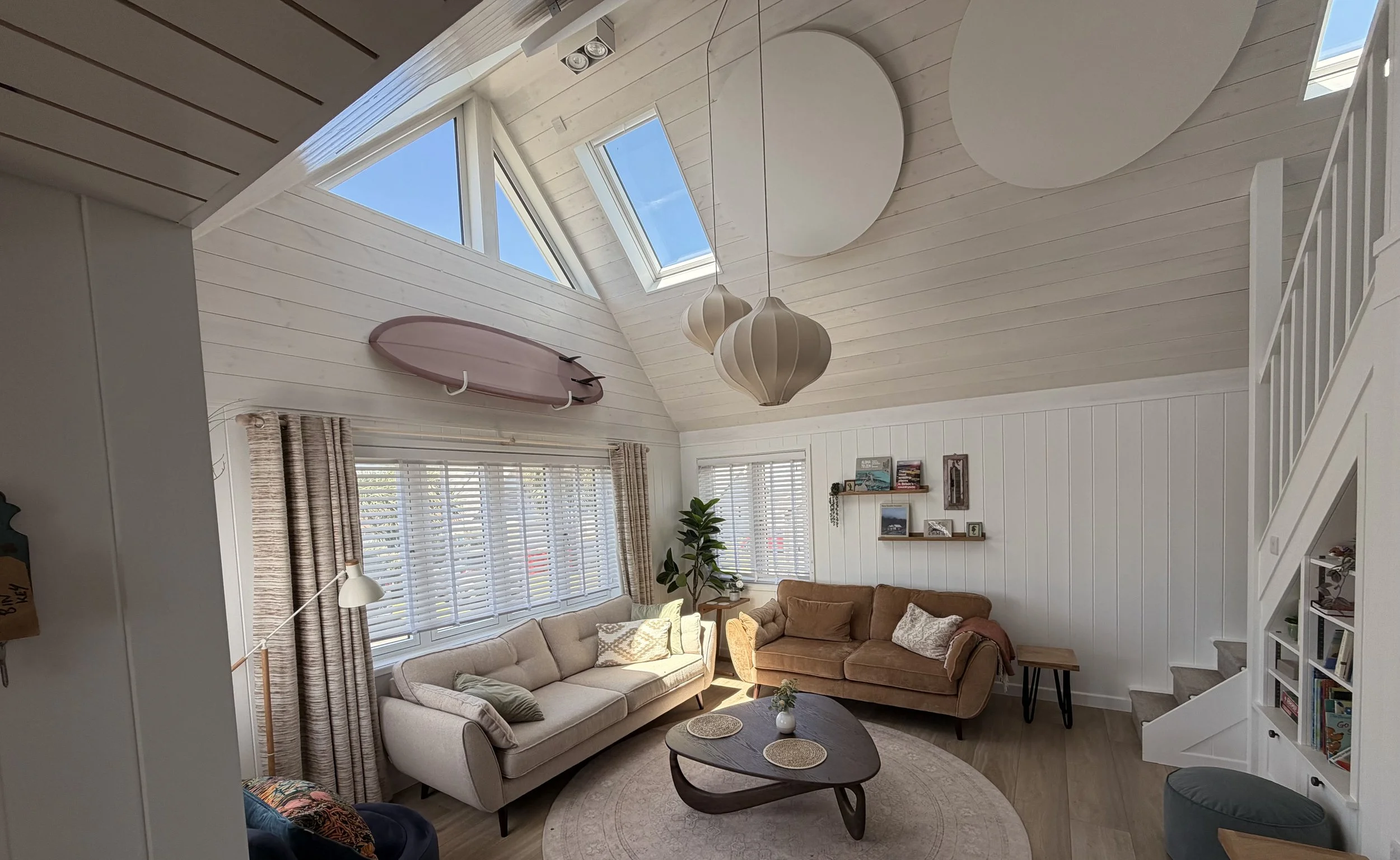 Living room with high wood-paneled ceiling, large windows with blinds, a pink surfboard mounted on the wall, and hanging pendant lights. The room has a beige sofa, a tan armchair, a wooden coffee table, and shelves with books and decor.