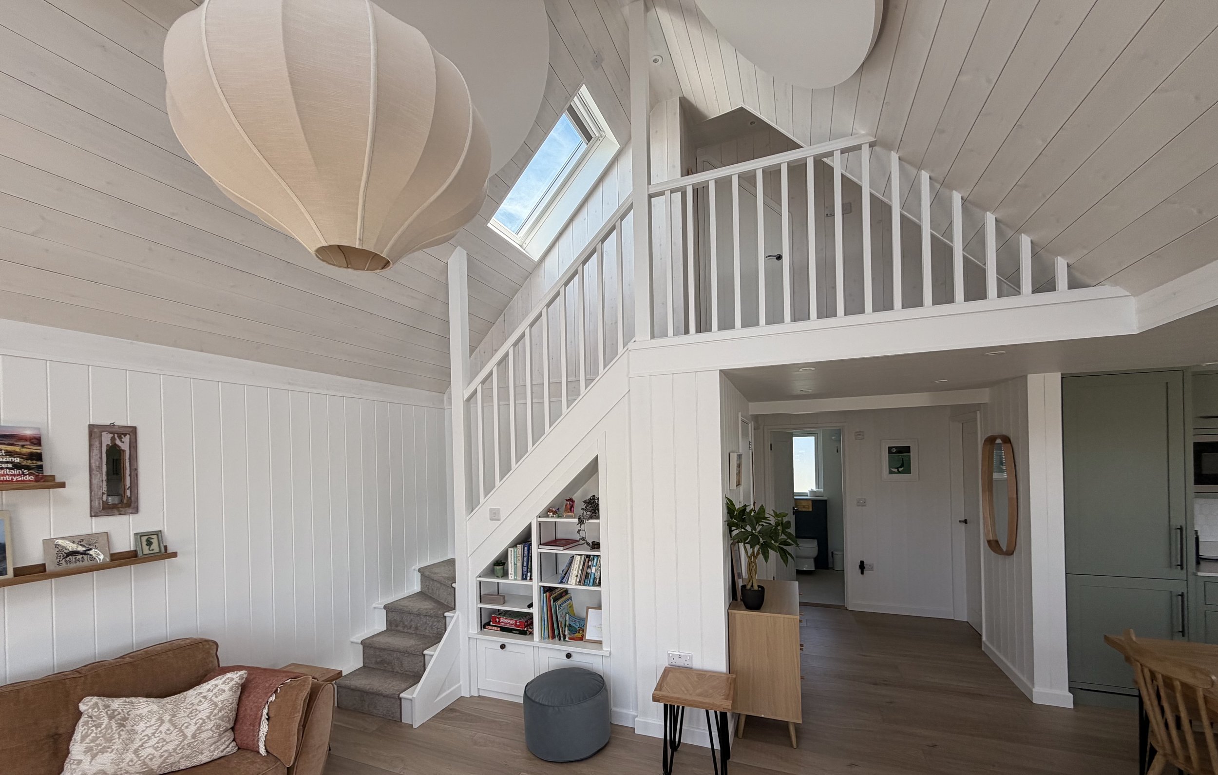 Living room with white wooden walls and ceiling, staircase leading to an upper floor, bookshelf, window and hanging lantern light fixture.
