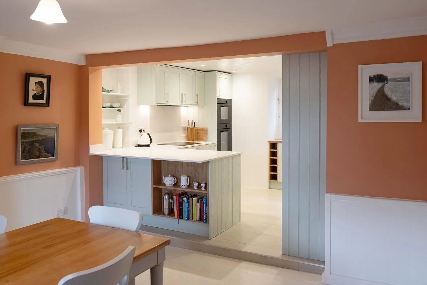 Open kitchen with white cabinets, a black oven, and a small white counter with open shelves holding books and teapots, visible from a dining area with a wooden table and white chairs.