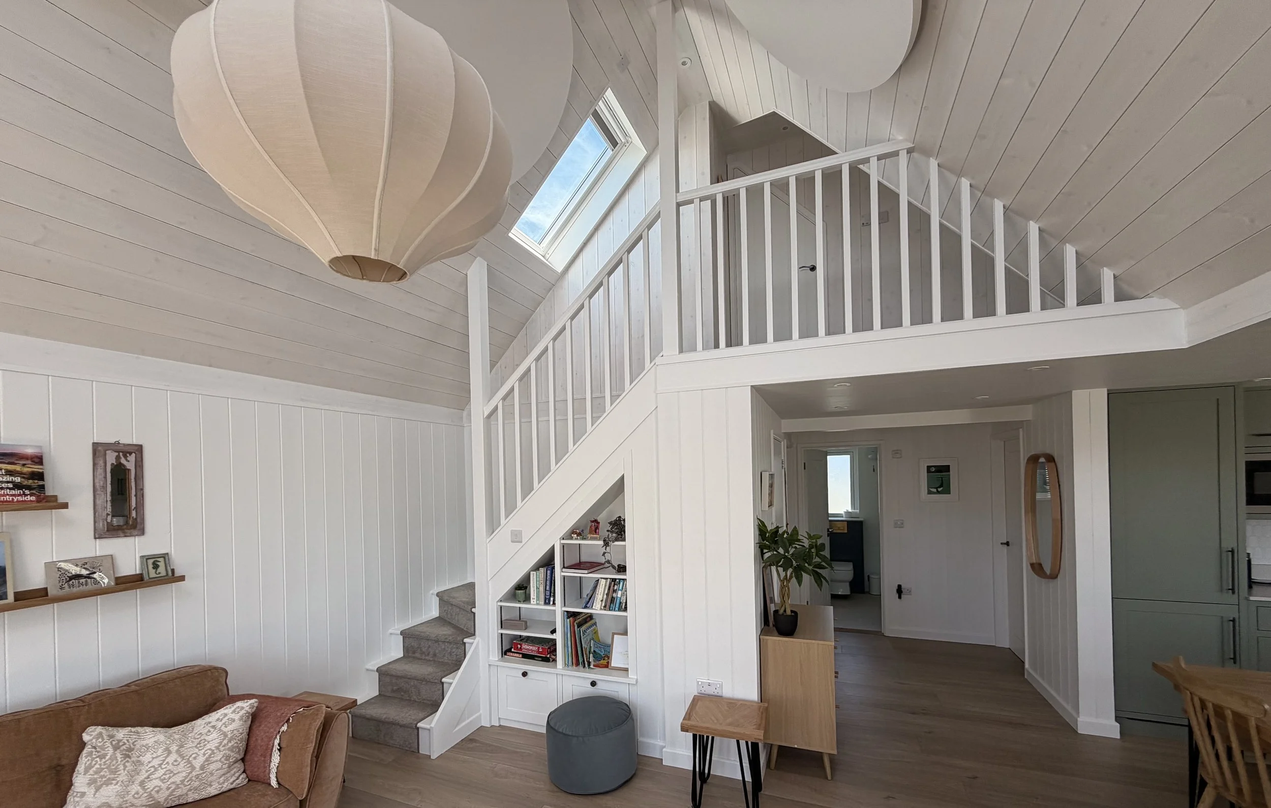Interior of a bright, modern living space with white walls, a staircase with white balustrades, a bookshelf filled with books, a window on the sloped ceiling, and a large hanging pendant light.