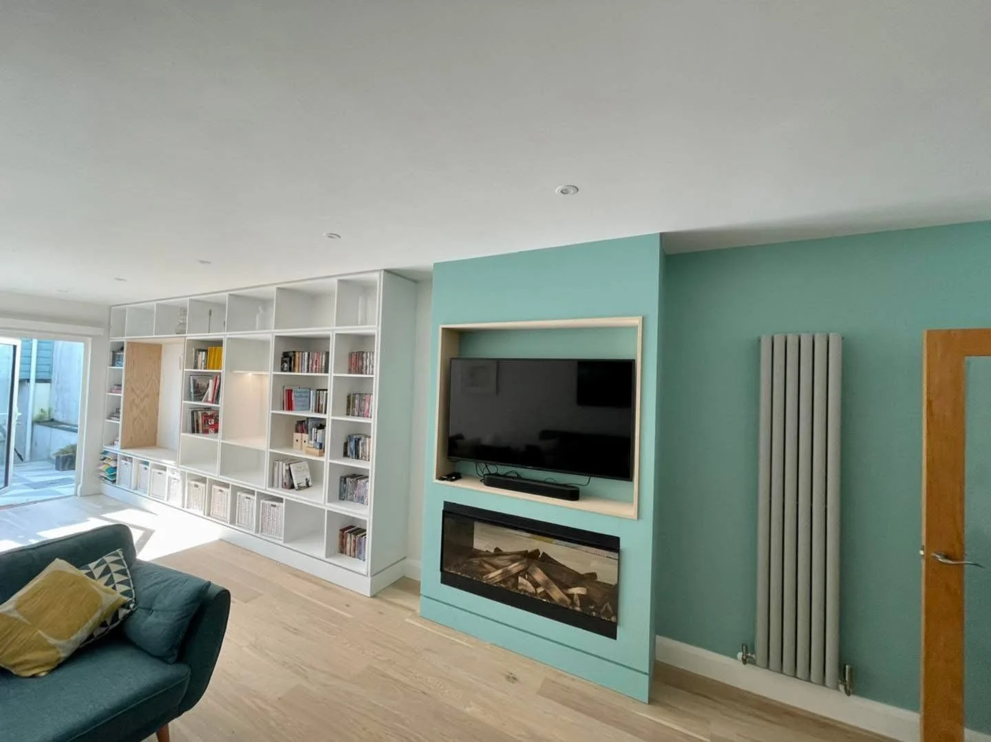 Modern living room with light-colored wood flooring, teal accent wall with a wall-mounted TV above an electric fireplace, and a large white bookshelf with books and decor. Sliding glass door leading to an outdoor patio is visible on the left.