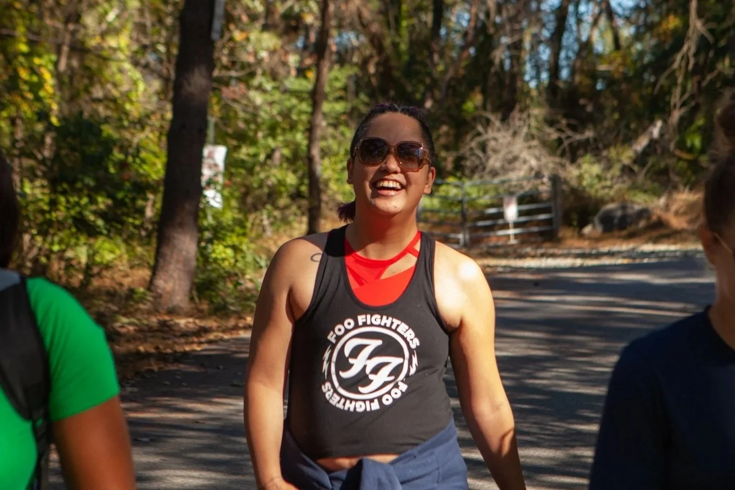 Woman smiling outdoors, wearing sunglasses and a Foo Fighters tank top, standing on a trail surrounded by trees.