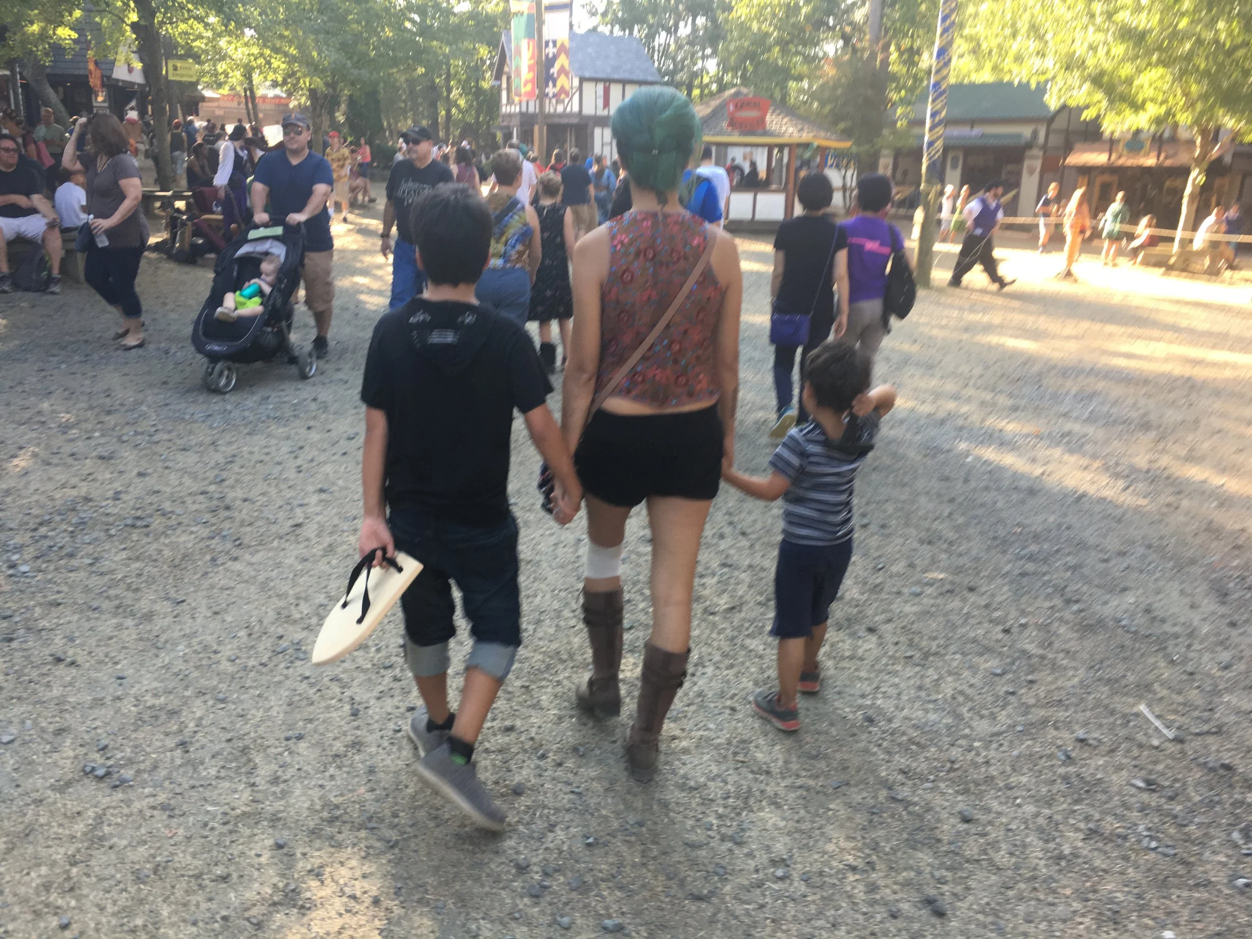 People walking in a park or festival with trees and booths in the background, seen from behind a woman with green hair holding hands with children.