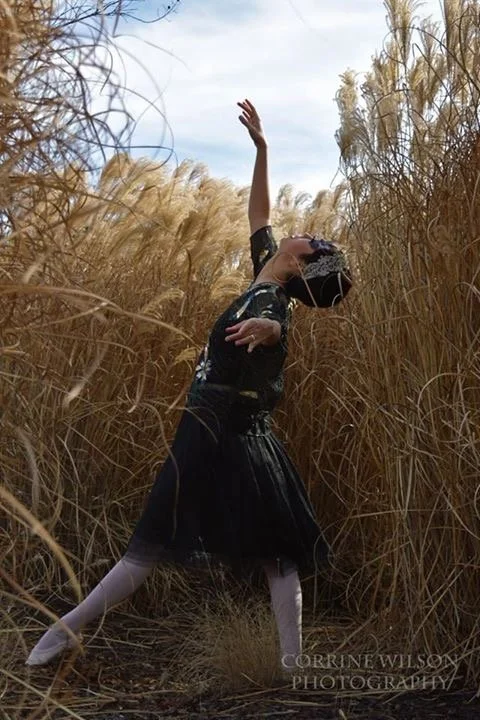 A girl in a black dress and white tights poses in a field of tall, dry grass with one arm raised and the other across her chest, wearing a masquerade mask.