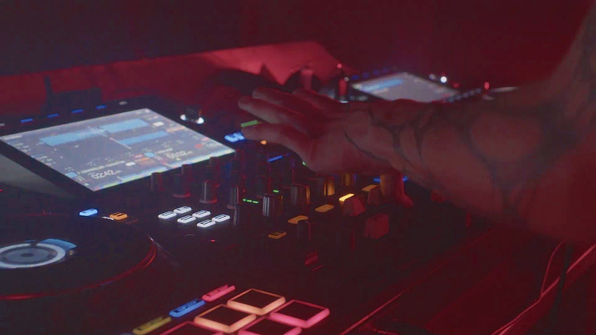 Close-up of a DJ's hand on a mixing console with colorful illuminated buttons under red and blue lighting.