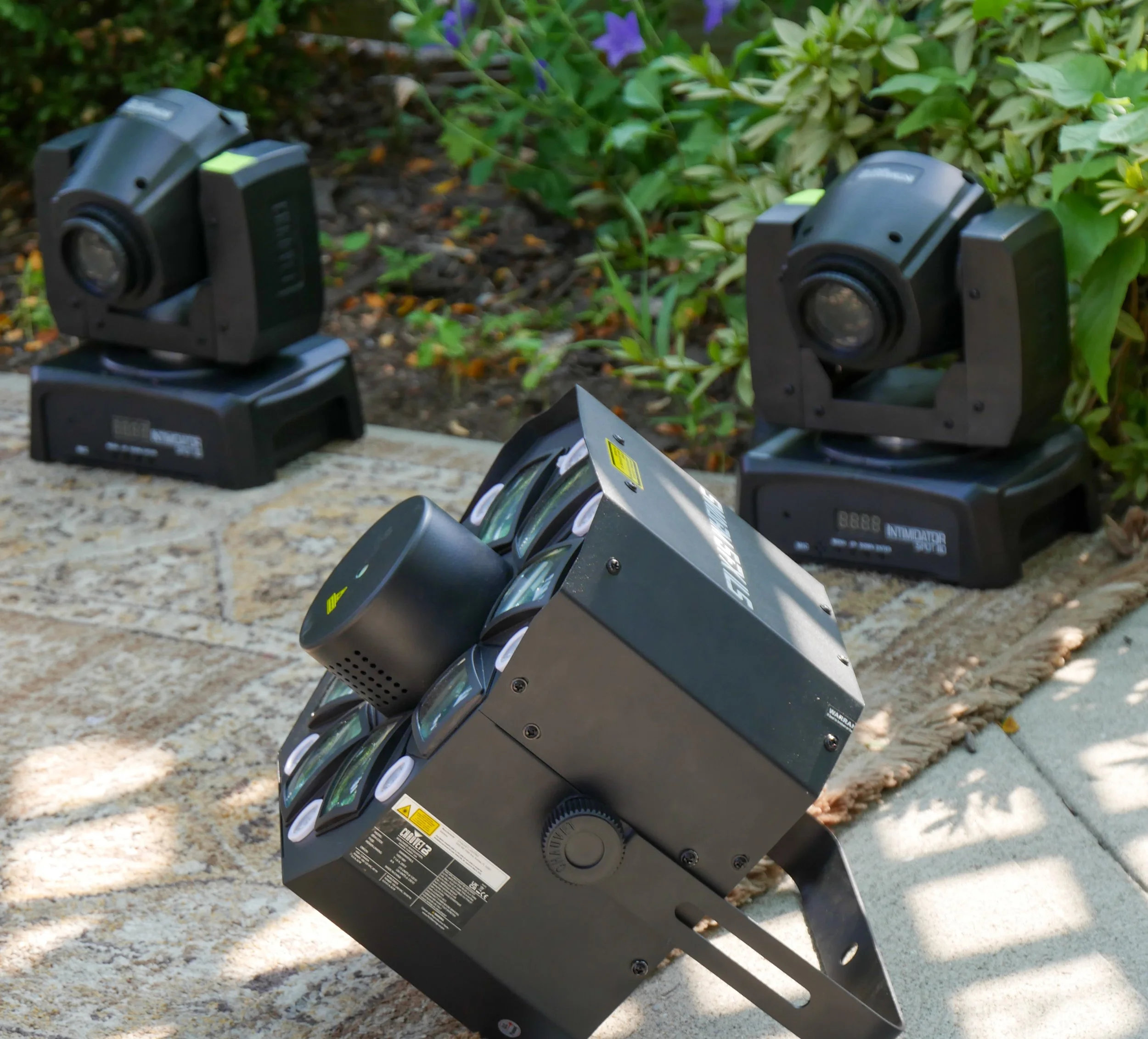 Several black outdoor lighting spotlights with adjustable heads mounted on a black structure, placed along a stone pathway with greenery in the background.