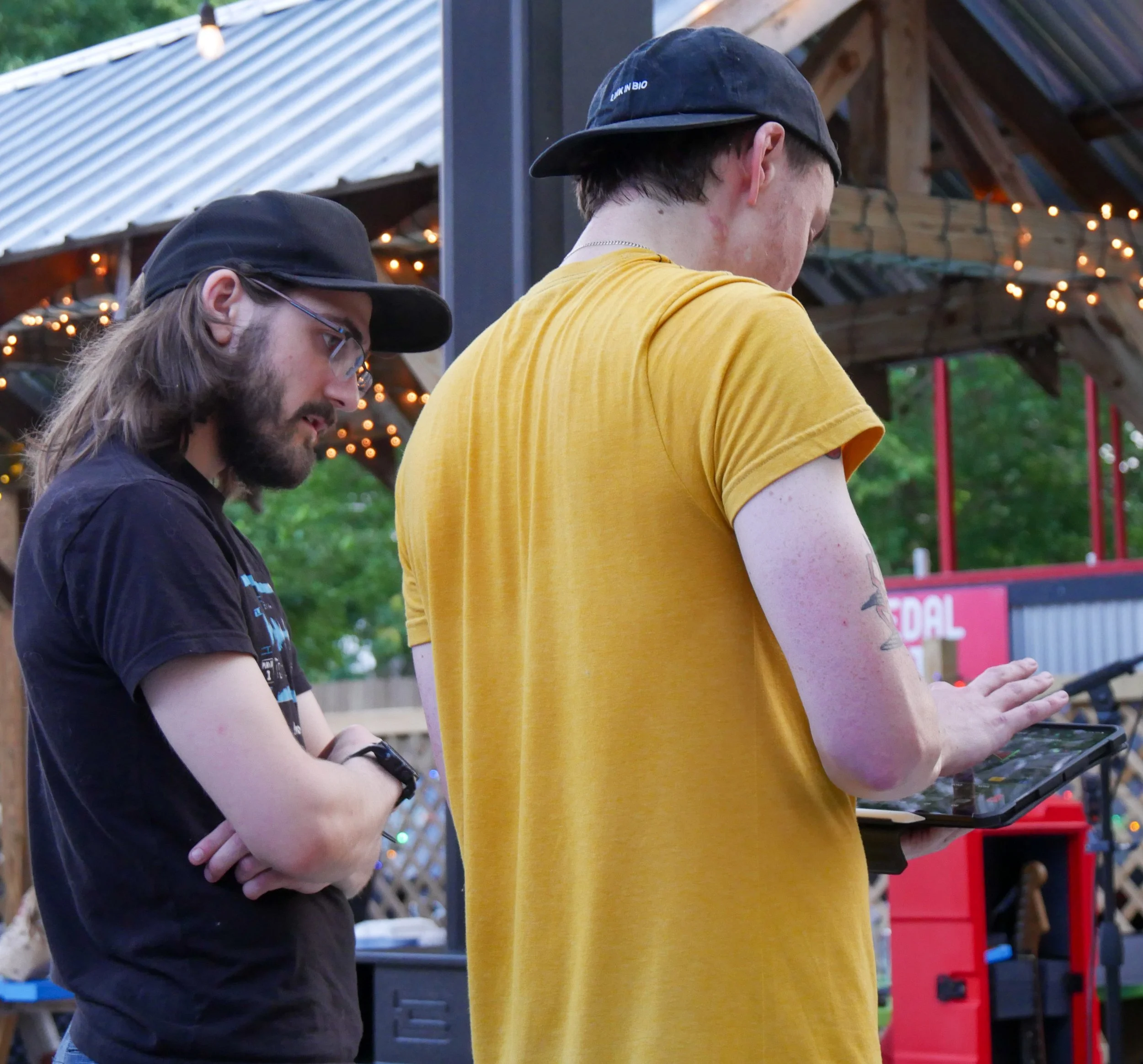 Two men standing outdoors, using smartphones. One man has long hair, glasses, and a beard, wearing a black cap and T-shirt. The other man has short hair, wearing a black cap and a yellow T-shirt. There are trees, a wooden structure, and string lights in the background.