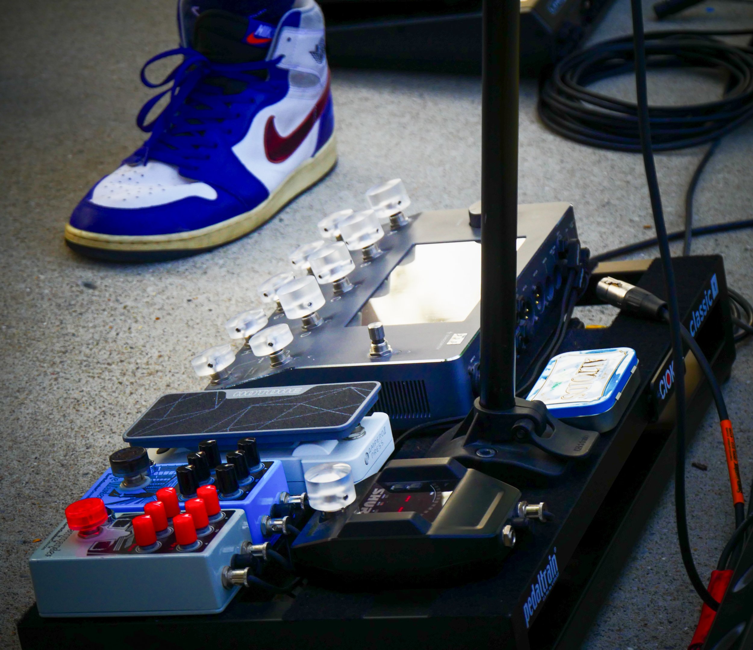 A collection of musical equipment and shoes on a gray carpeted floor, including a pair of Nike high-top sneakers, electronic pedals, cables, and a smartphone.