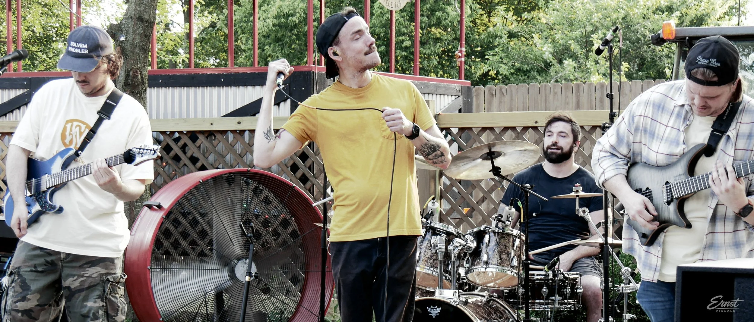 A band performing outdoors with four members: a guitarist wearing a white shirt and camouflage pants, a vocalist in a yellow shirt holding a microphone, a drummer with a beard playing drums, and another guitarist wearing a plaid shirt. The background has trees, a wooden fence, and metal fencing.