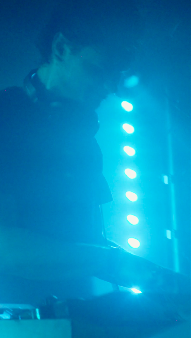 A person standing in a dark room illuminated by blue stage lights, with a guitar.