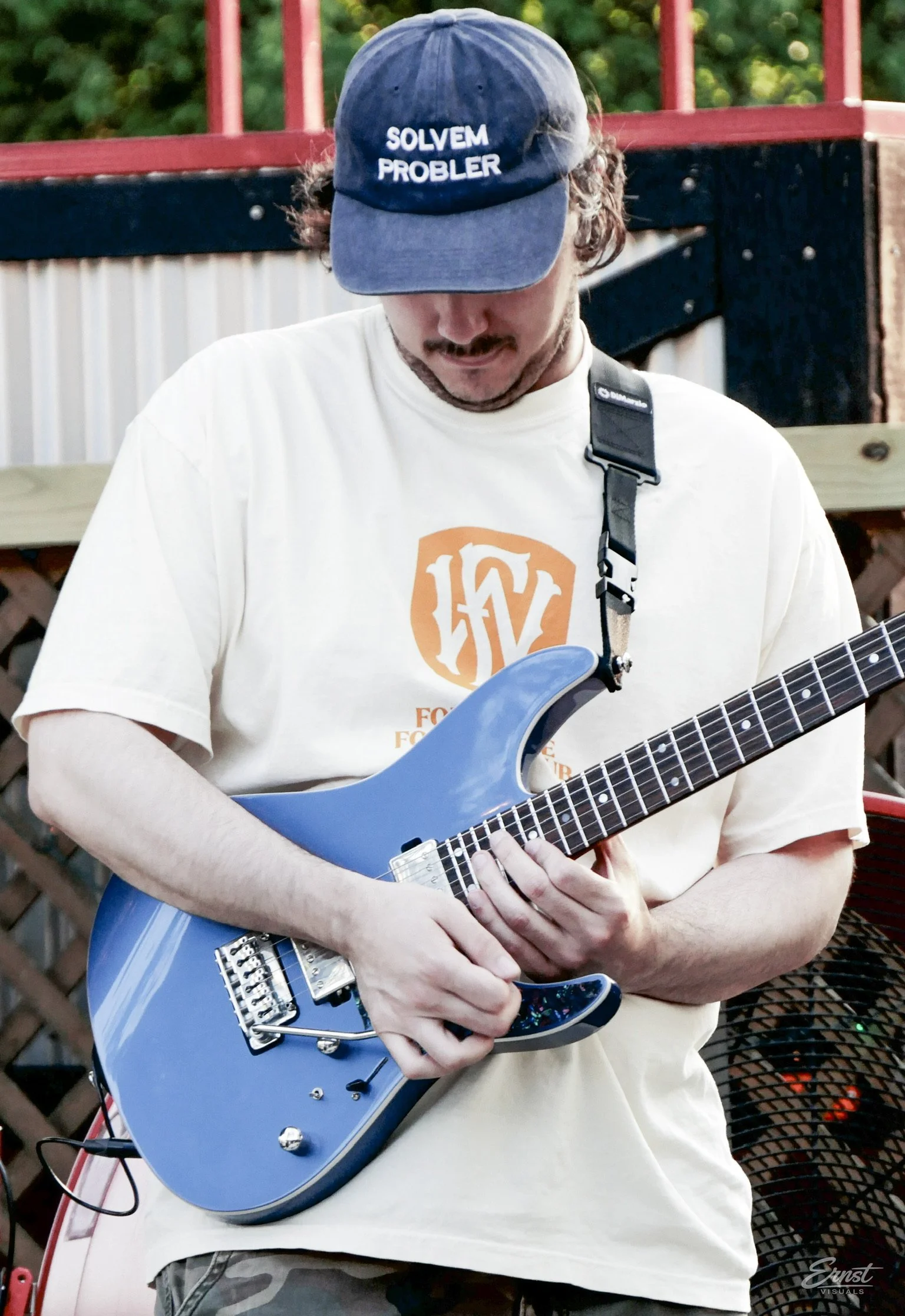 A man with curly hair wearing a baseball cap with the words 'SOLVEM PROBLER' on it, playing a blue electric guitar. He is dressed in a cream-colored t-shirt with an orange logo and is outdoors with greenery in the background.