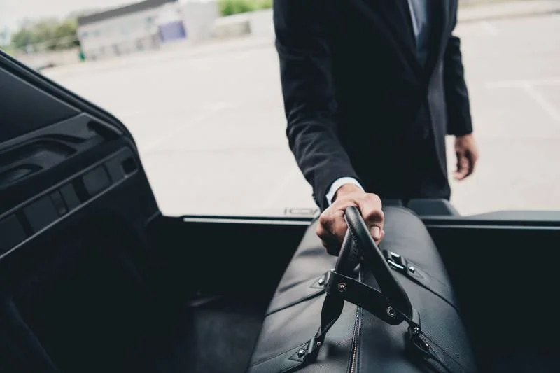 Person in a black suit holding a black briefcase outside of a vehicle in a parking lot.