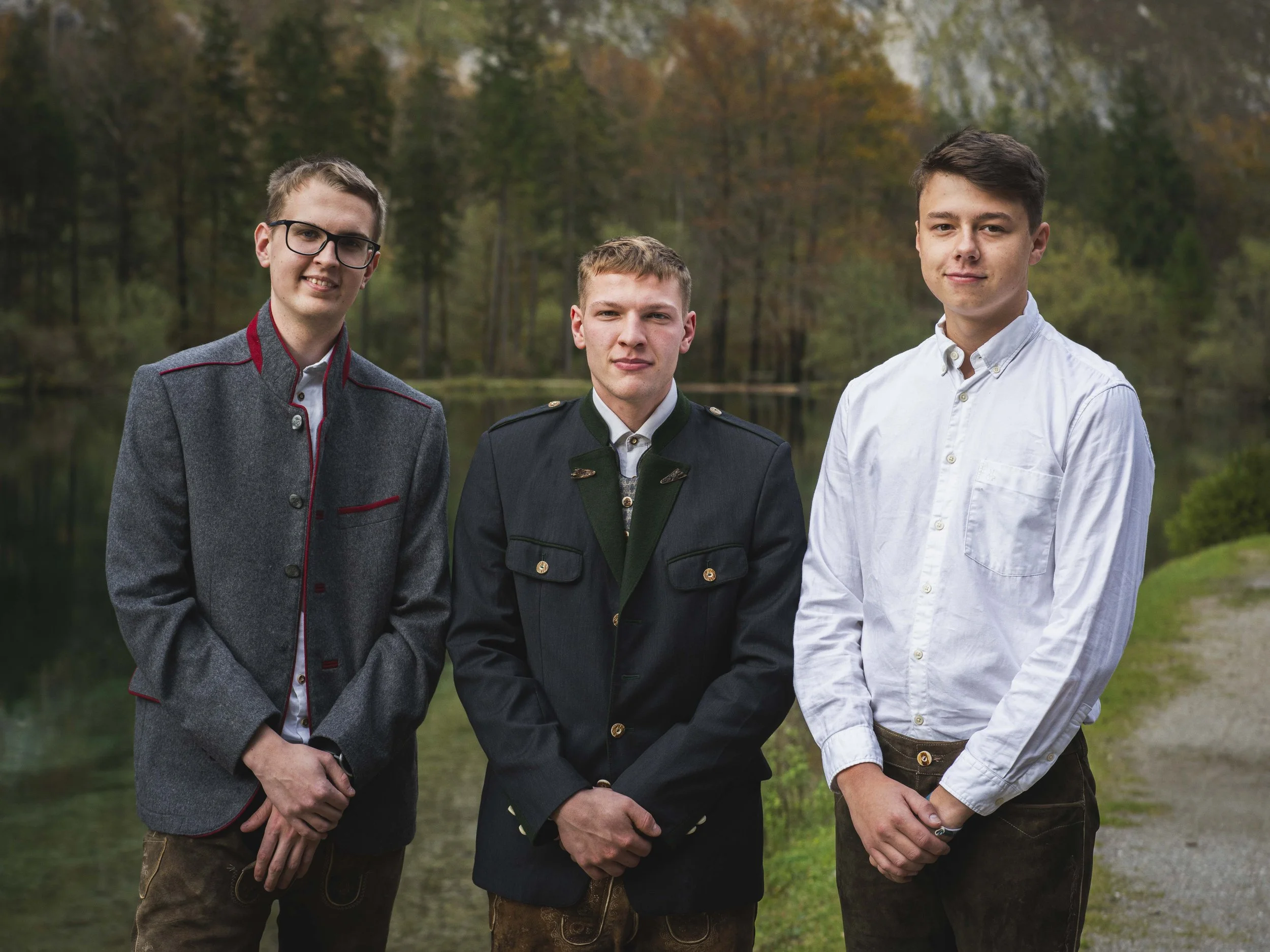 Drei junge Männer stehen am Ufer eines Sees mit Bäumen im Herbst im Hintergrund. Zwei Männer tragen traditionelle Trachtenjacken, der dritte trägt ein weißes Hemd.