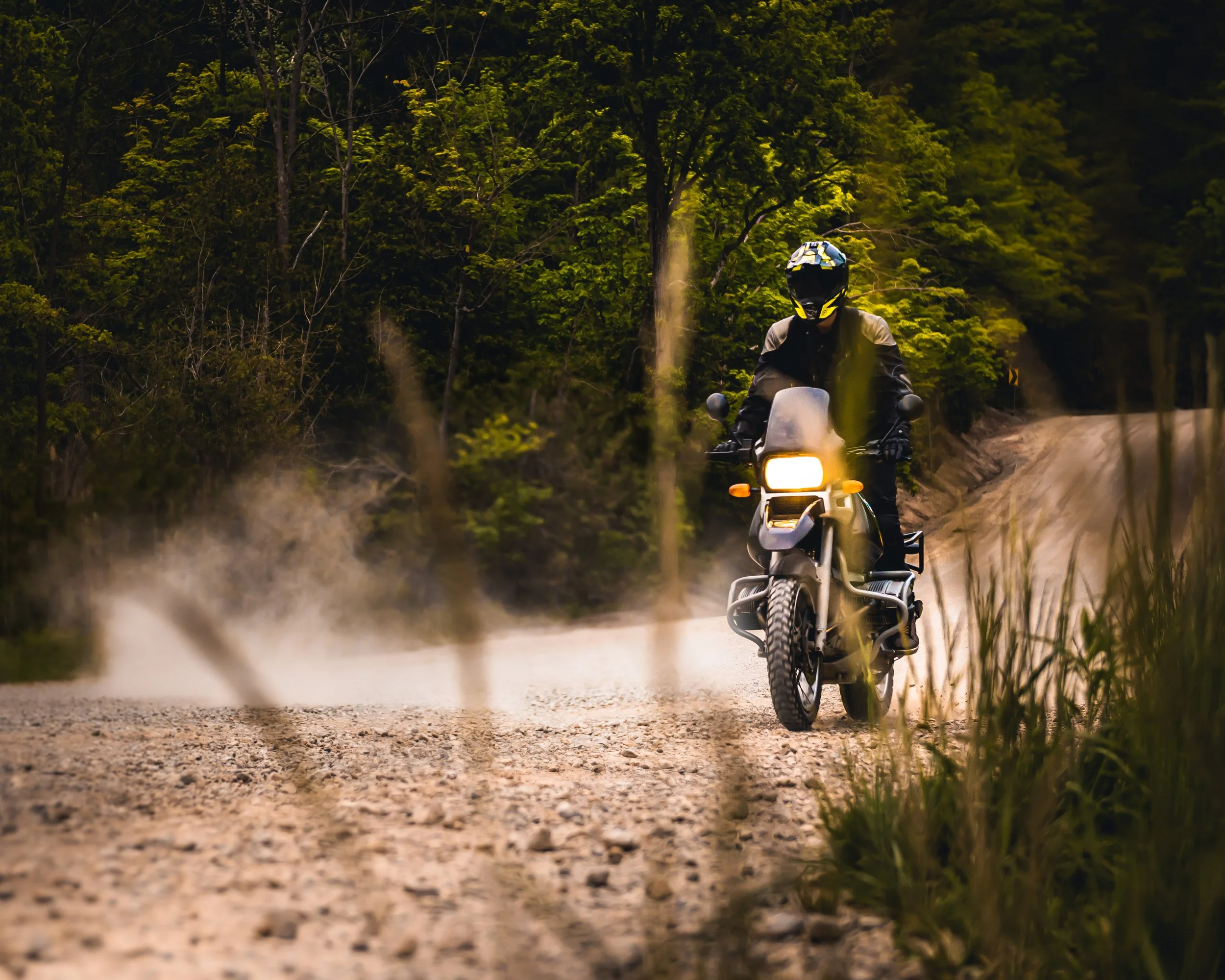 A person riding a motorcycle along a dirt road surrounded by lush green trees, seen from a low angle through grass and plants.