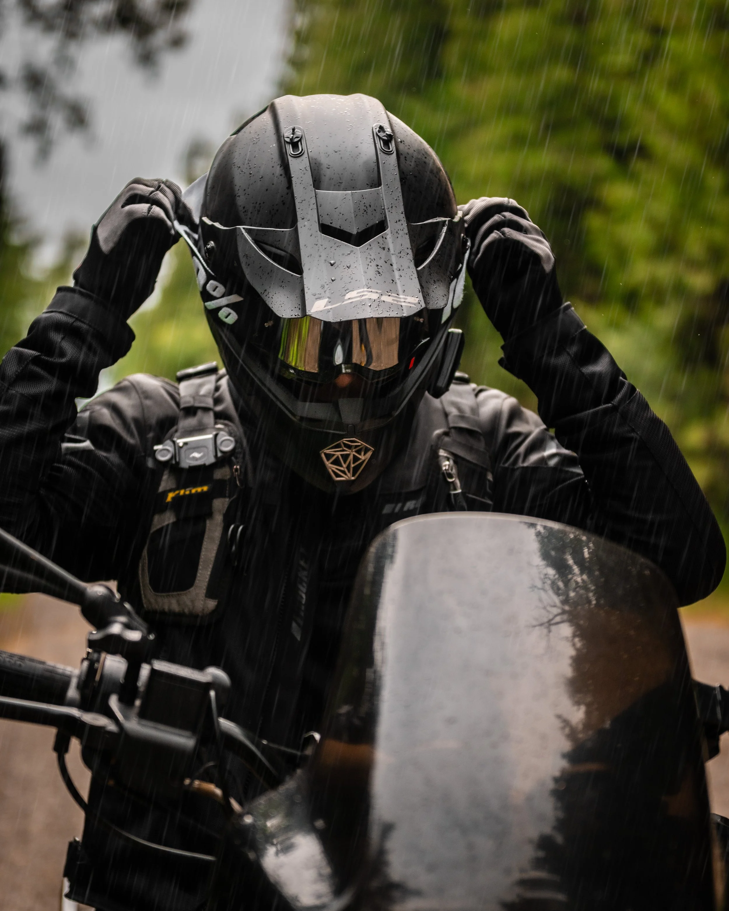 A person wearing a motorcycle helmet and black gear riding a motorcycle in the rain, with trees and a cloudy sky in the background.