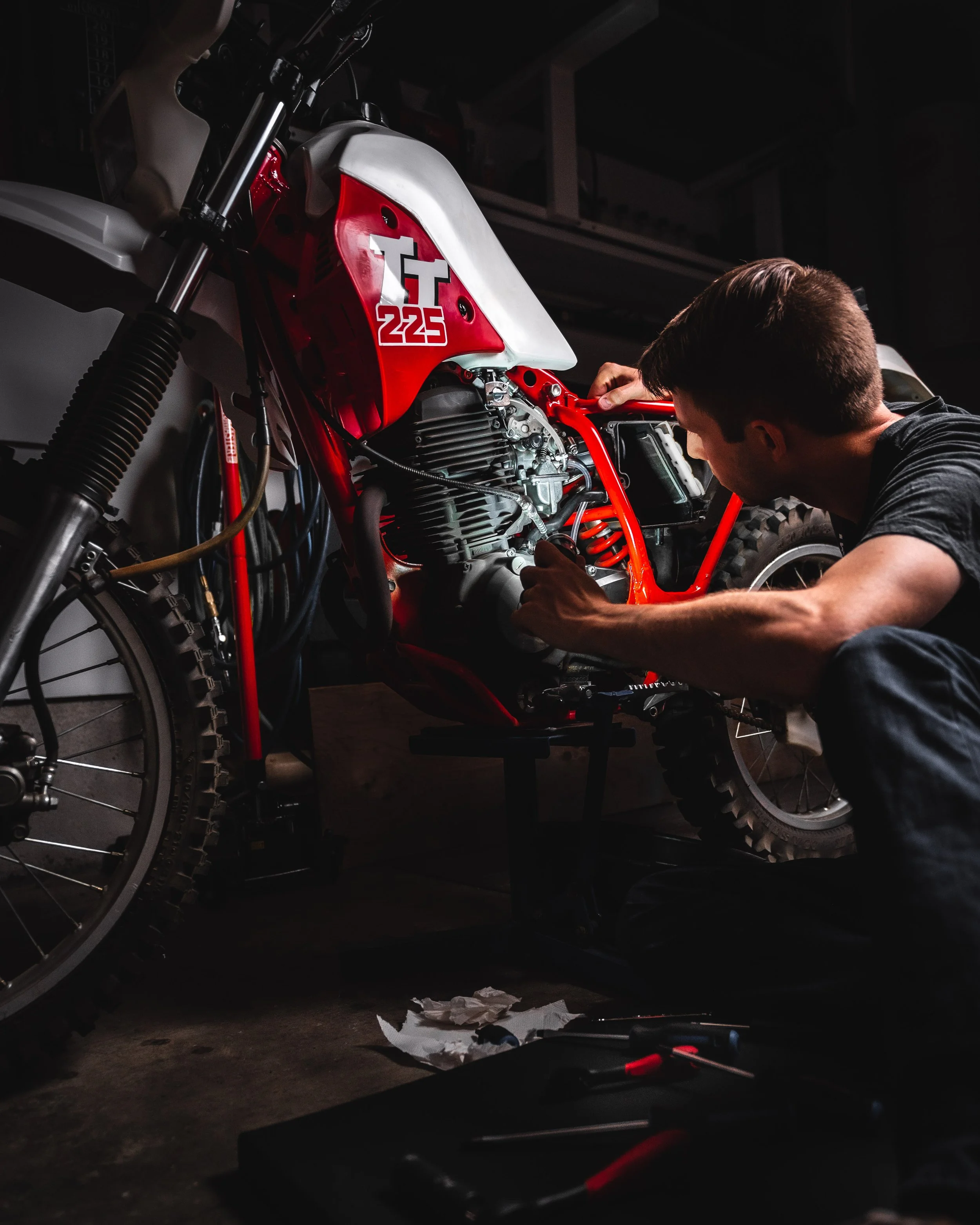 A person working on a red and white motorcycle in a dark garage. The motorcycle has a sticker that reads 'TT 225'. The person appears to be tightening or adjusting parts around the engine area. There are tools and paper on the floor.