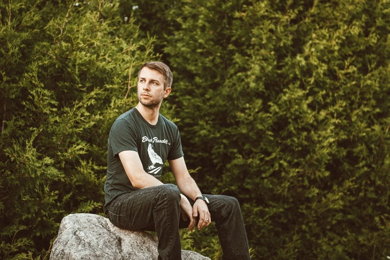 A man sitting on a rock outdoors, surrounded by green foliage, looking to the side.