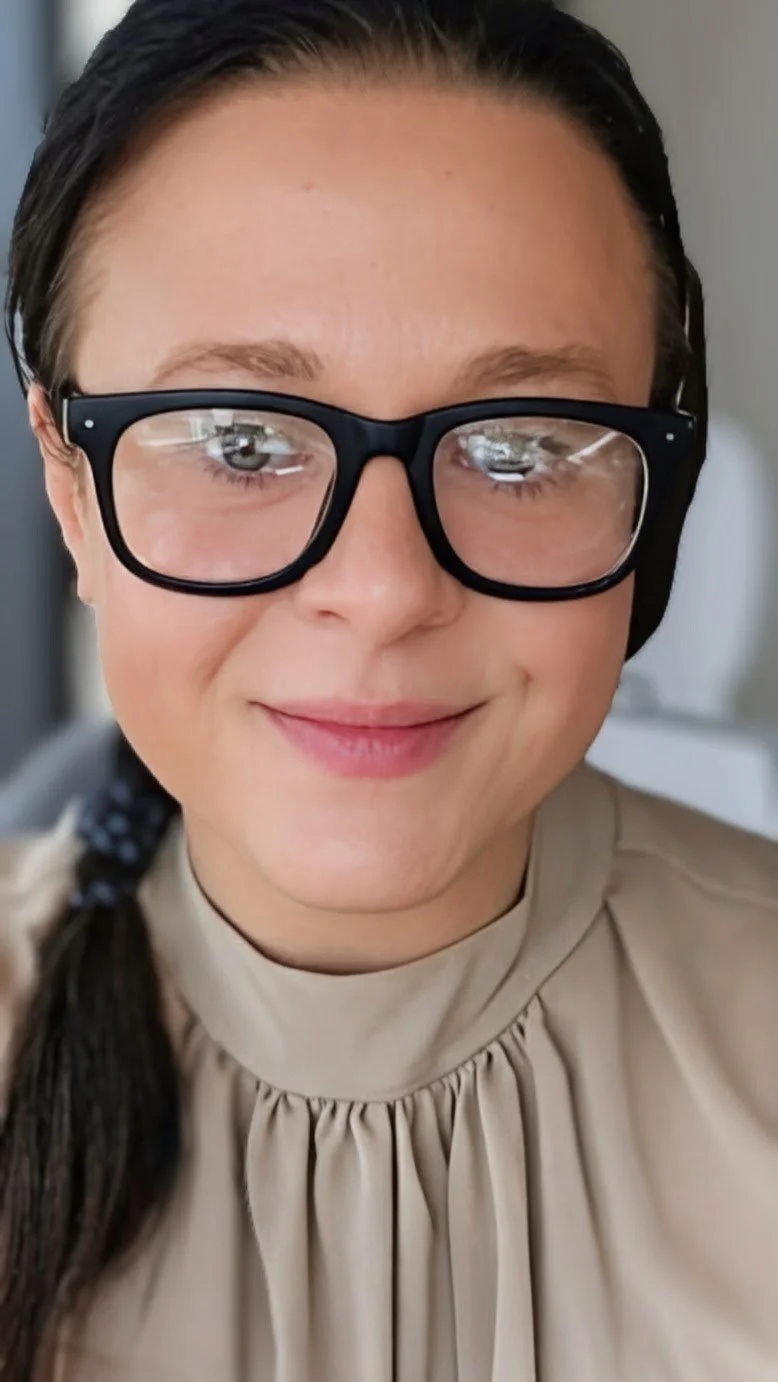 A close-up of a woman with dark hair, wearing glasses and a beige, high-collared blouse. She has a small smile and is looking directly at the camera.