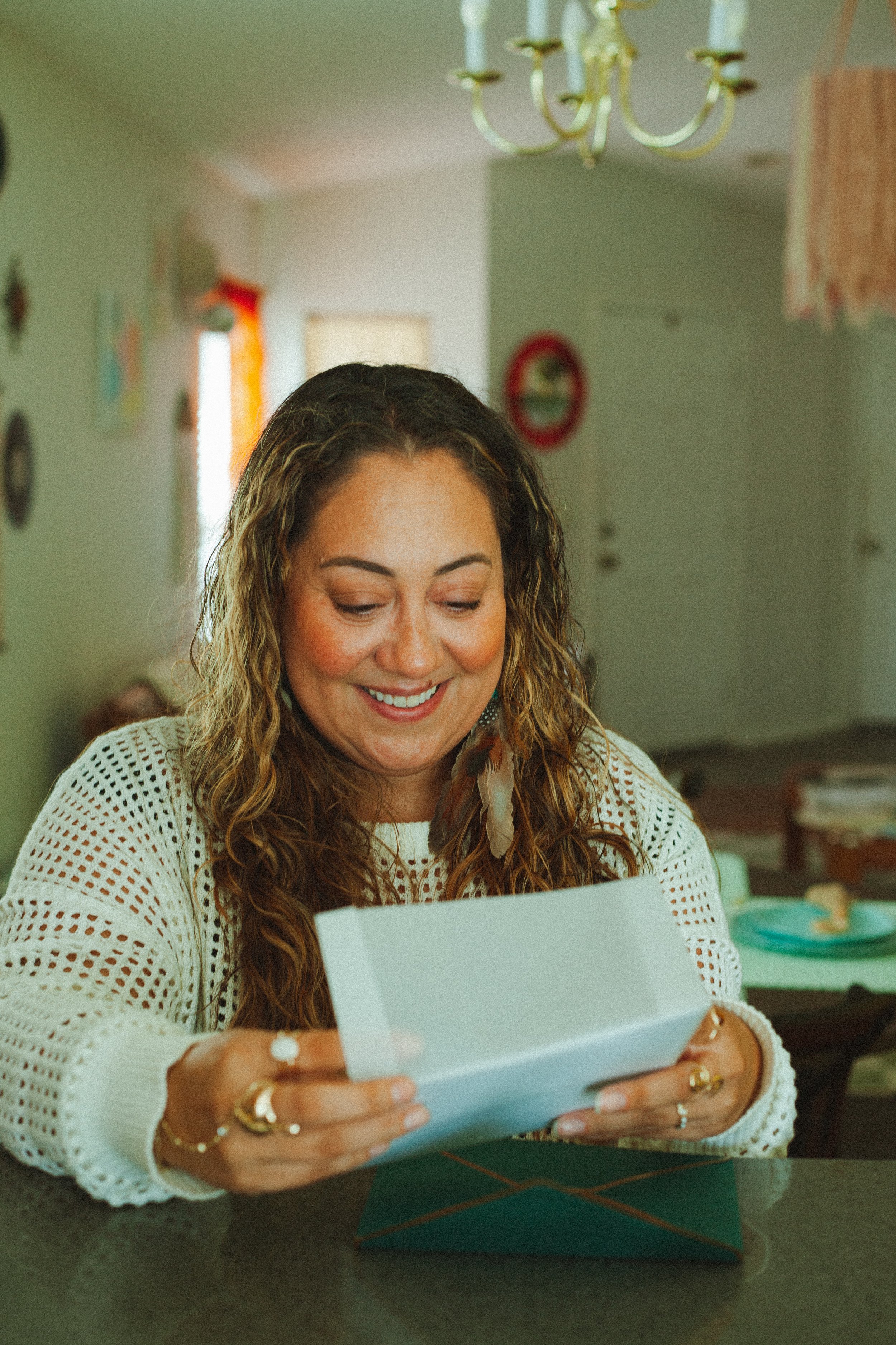 A woman with curly hair, wearing a white open-knit sweater, smiling as she looks at a greeting card, sitting at a table in a brightly decorated room.