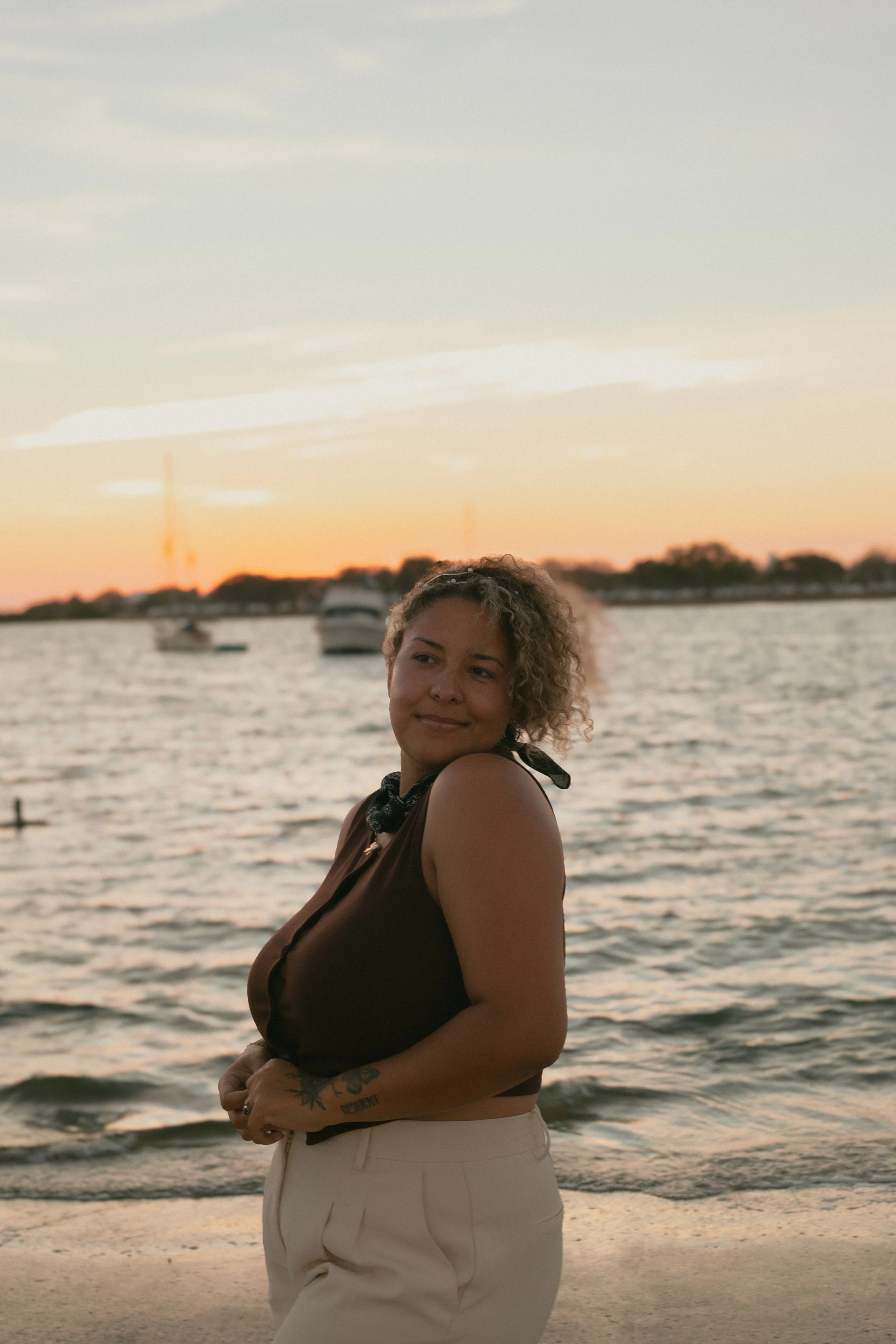 A woman with curly hair stands on a beach at sunset, facing the water with boats in the background.