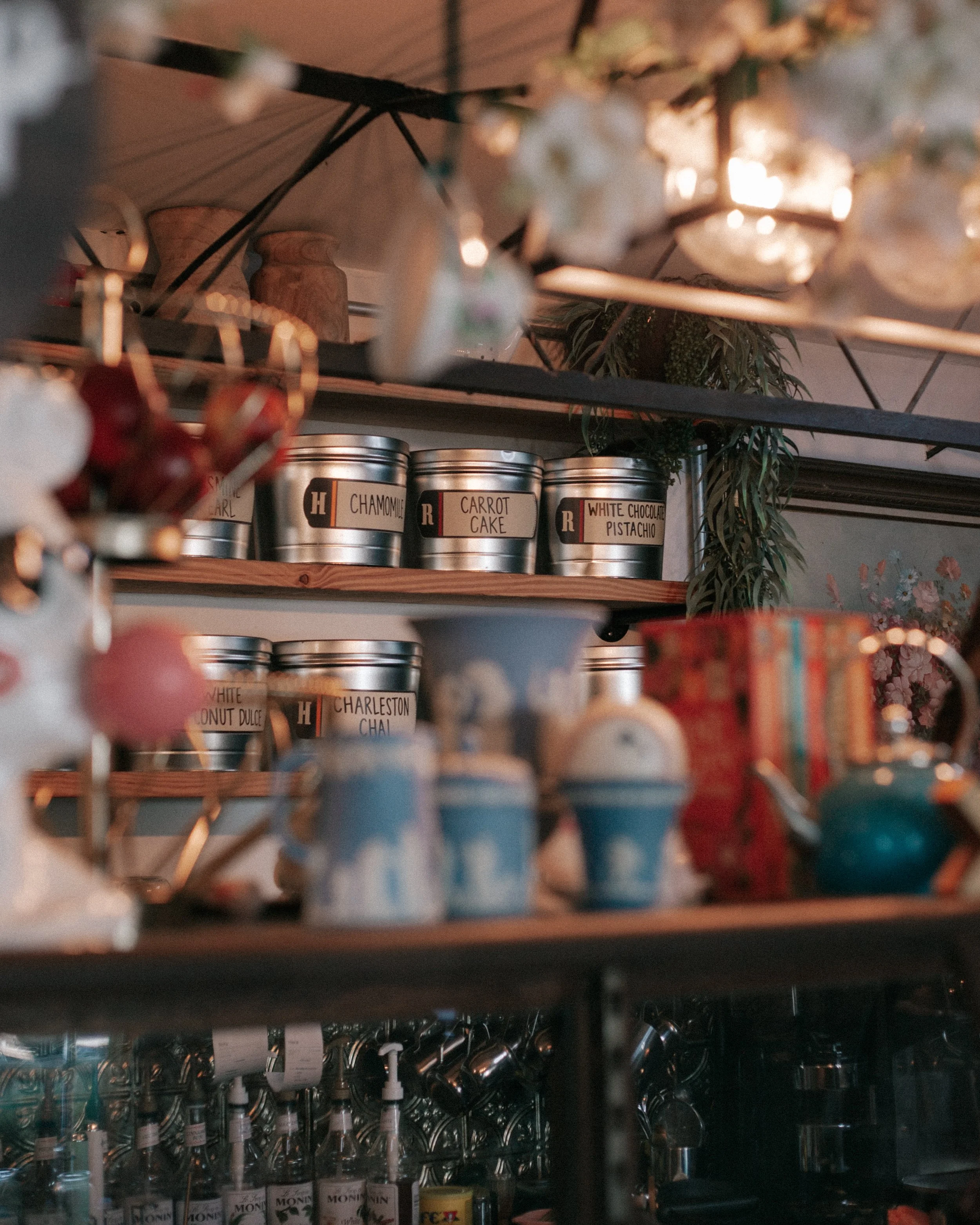 Shelf with metal containers labeled with flavors such as chamomile, carrot cake, white chocolate pistachio, and others, in a kitchen or store setting.
