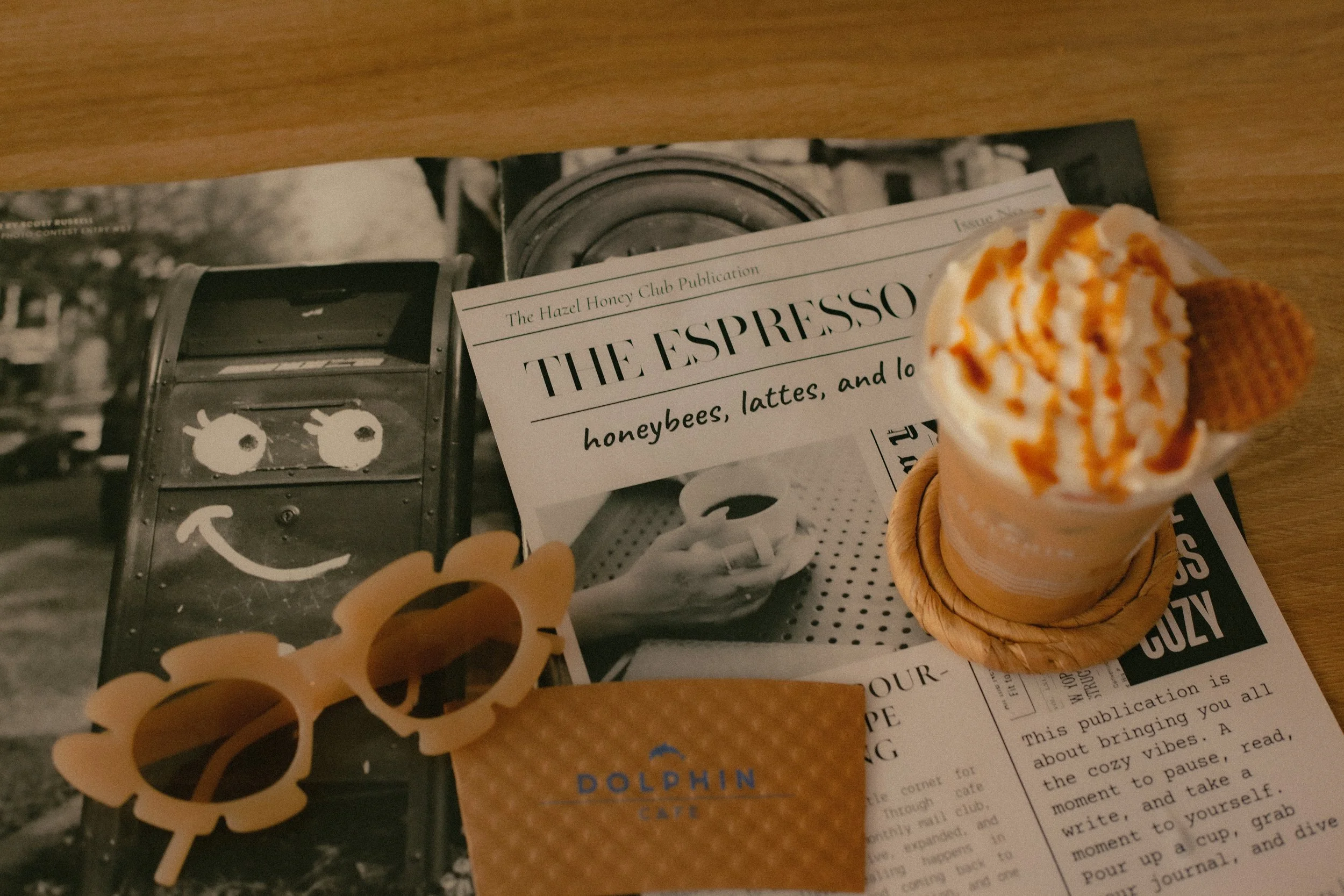 A table with a black and white magazine, a printed menu, a scoop of ice cream with caramel drizzle in a waffle cone, and orange sunglasses.