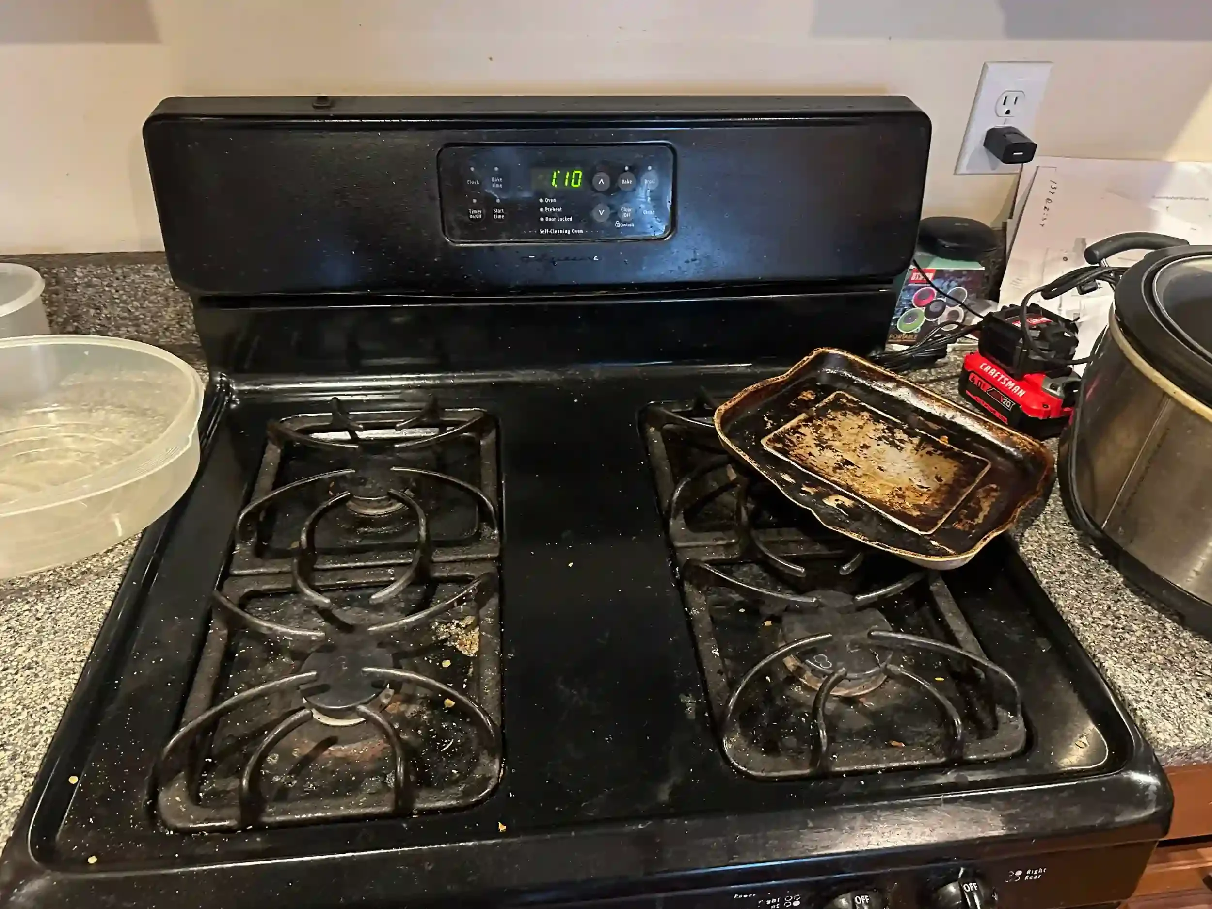 A black stove with burners and control knobs, a dirty baking sheet, and a used stove top with food debris, next to a container with a clear plastic lid and a paper bag.