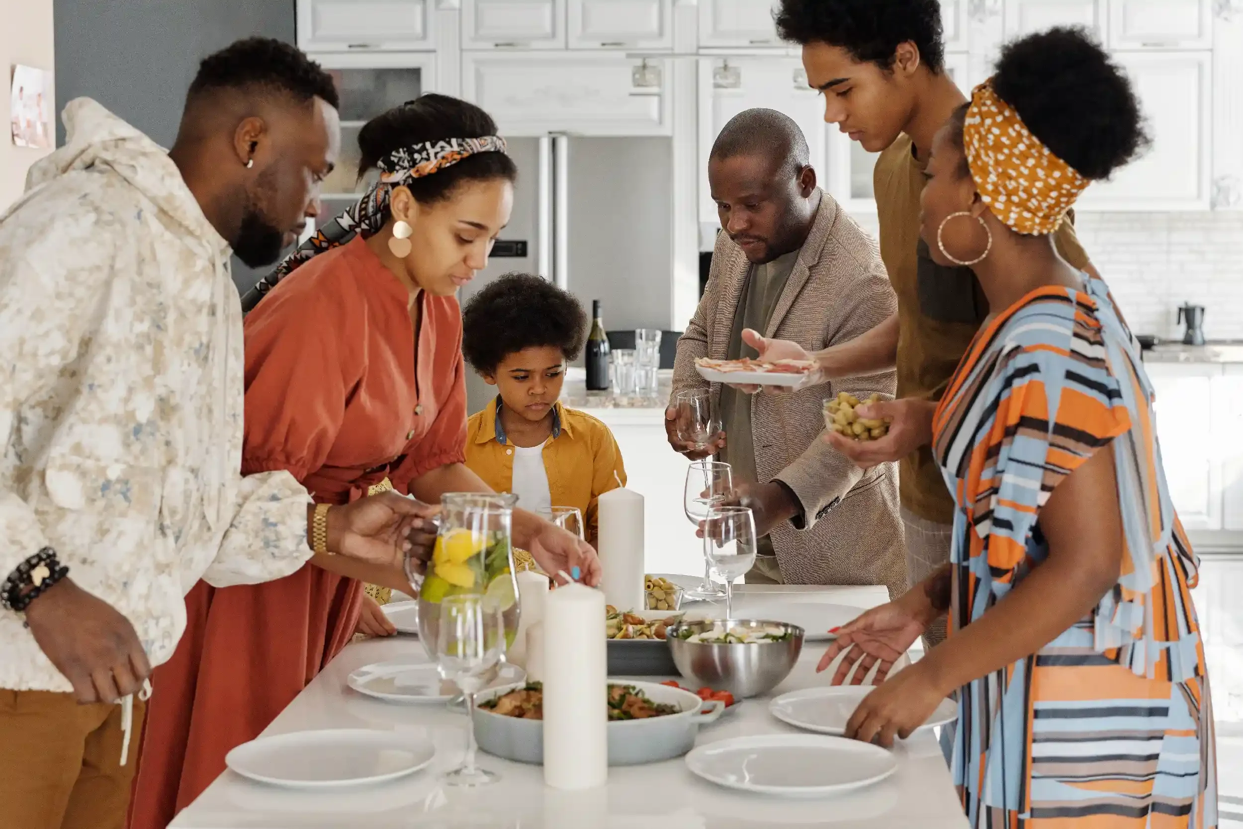 A diverse group of six adults and one child gathered around a table in a kitchen, serving and preparing food for a meal.
