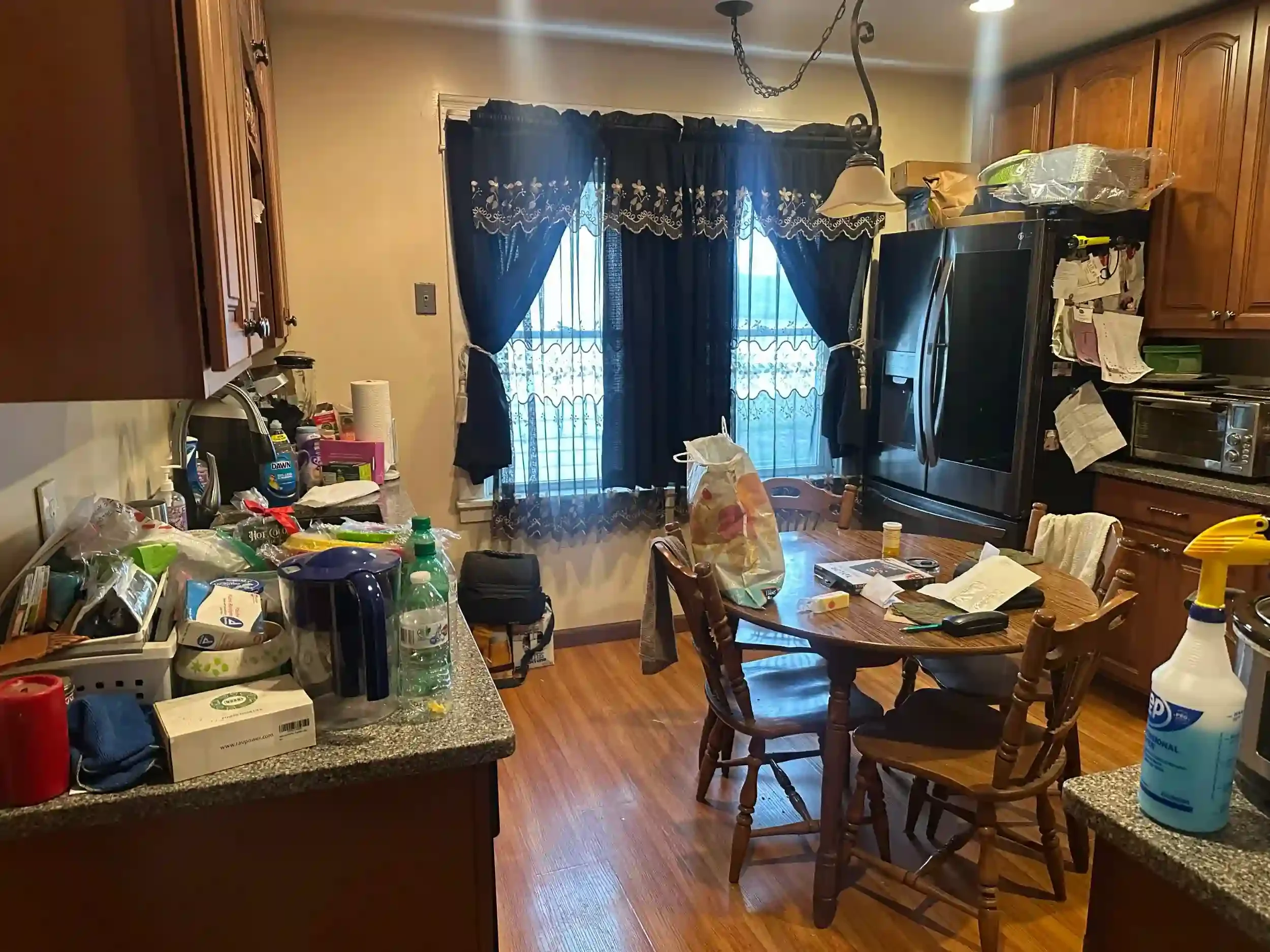 A cluttered kitchen and dining area with dark curtains, wooden cabinets, a microwave, and a table with various items including food, cleaning supplies, and electronics.