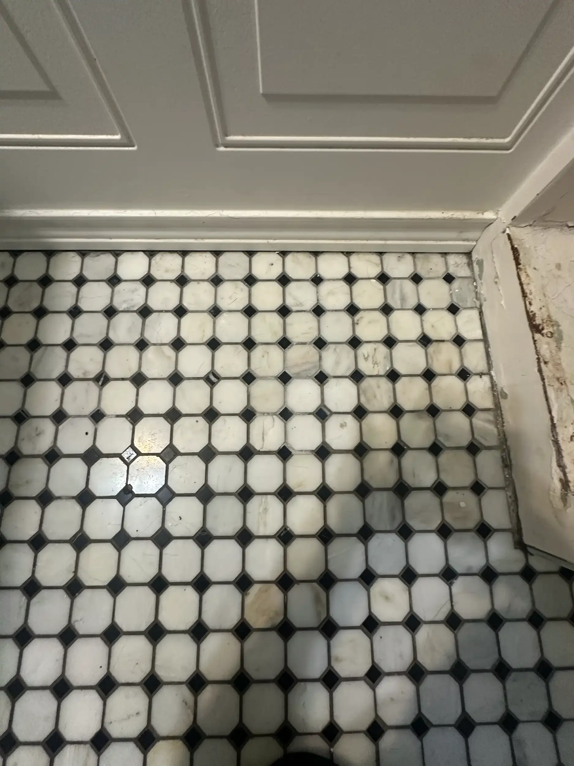 Close-up of a tiled floor at the base of a gray door and wall in a bathroom or kitchen. The tiles are hexagonal, with some showing signs of wear and stains.