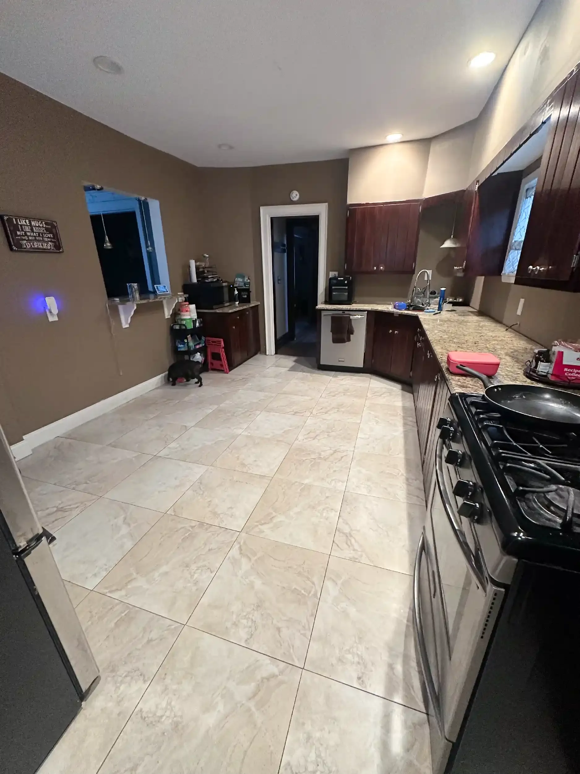 A kitchen with tiled floor, dark wood cabinets, granite countertop, and a stove. There is a bowl on the stove, and some kitchen items on the counter. An open door leads to another room, and a small black dog is near the wall below a window. There is 