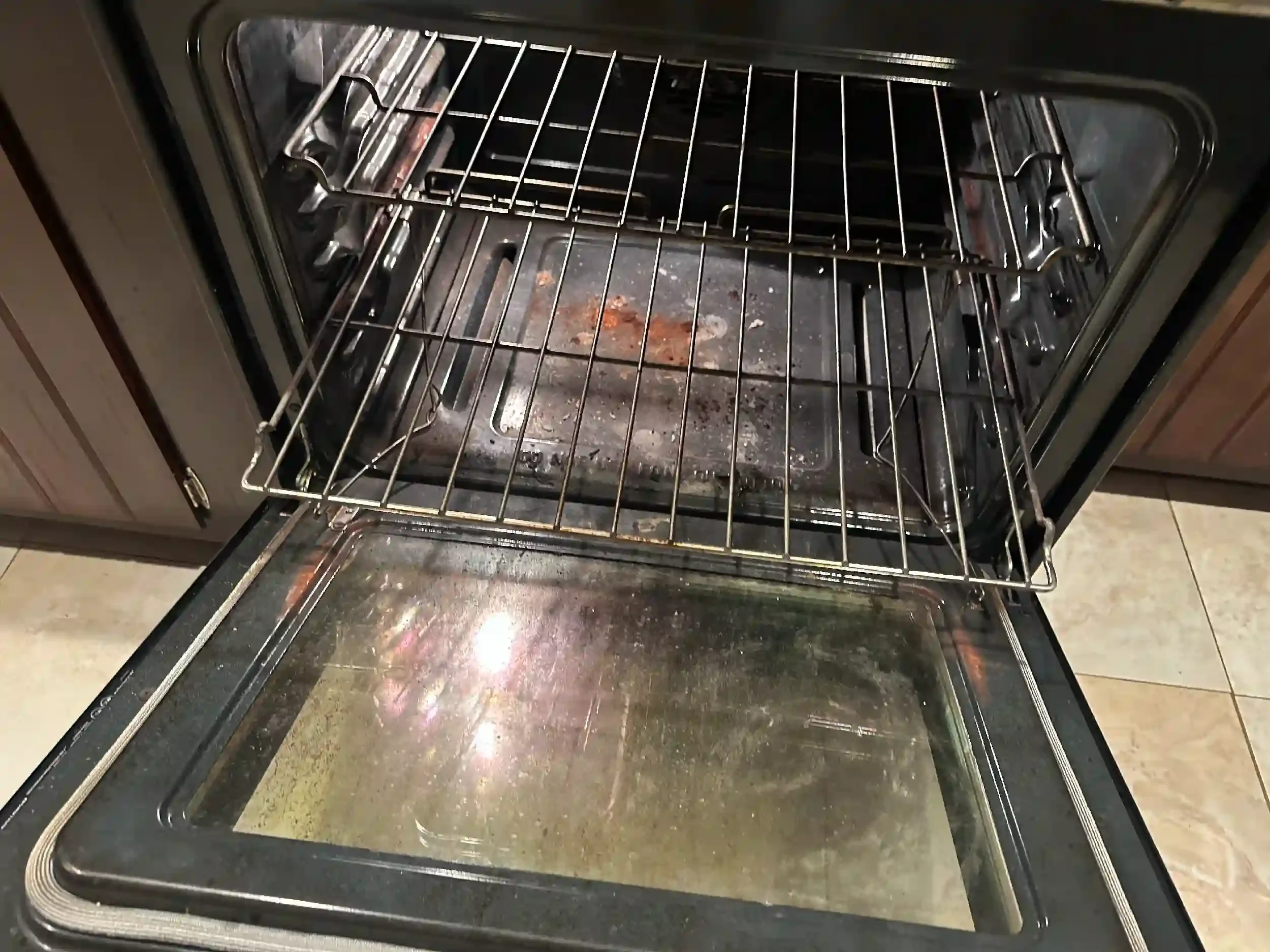 Empty oven with some burnt food residue and grime on the oven floor and racks, with the oven door open.