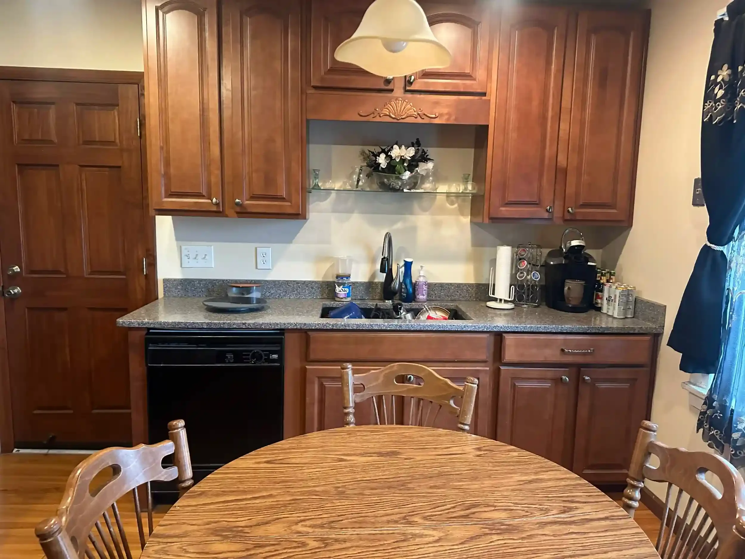View of a kitchen with wooden cabinets, a countertop with various items, a sink, a dining table with chairs, and a hanging light fixture. There are kitchen appliances and decorative items on the counter.