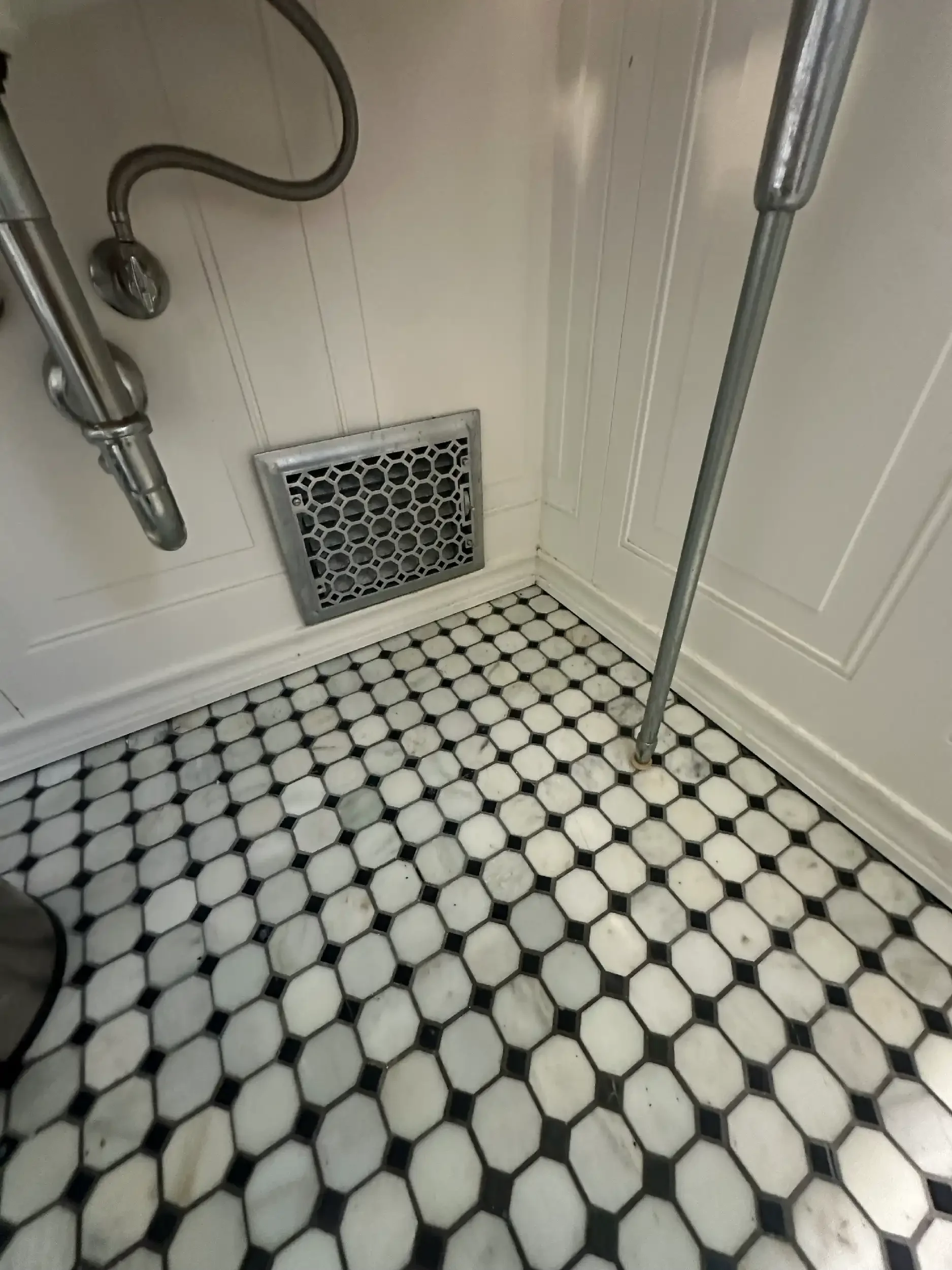 Close-up of a bathroom floor with white hexagonal tiles with black accents, a floor register, and a section of a wall with white wainscoting and corner molding. There is a metal shower pipe and a grab bar in the shower area.