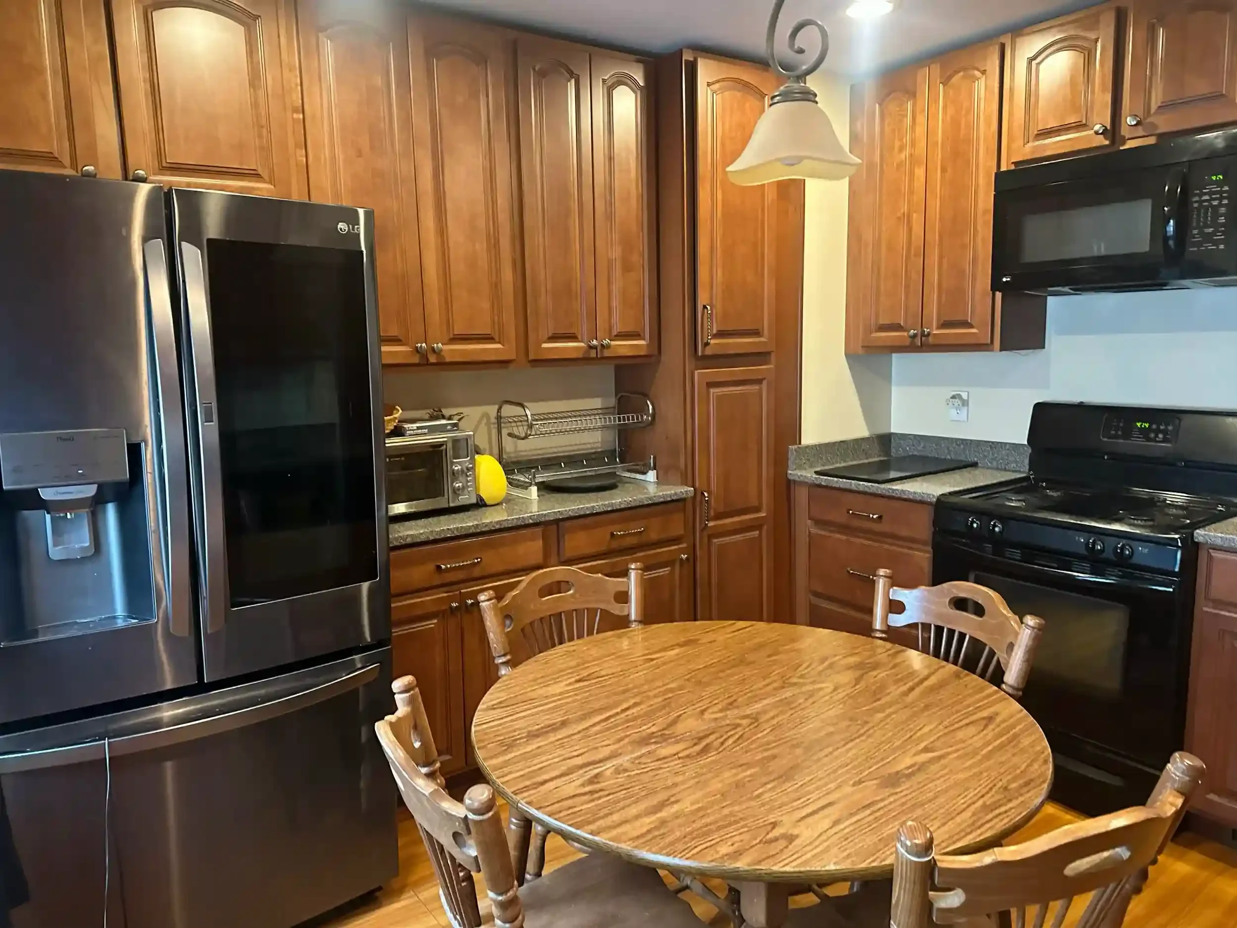 Kitchen with wooden cabinets, a stainless steel refrigerator, a black microwave, a toaster oven, a gas stove, a hanging lamp, and a round wooden table with chairs.