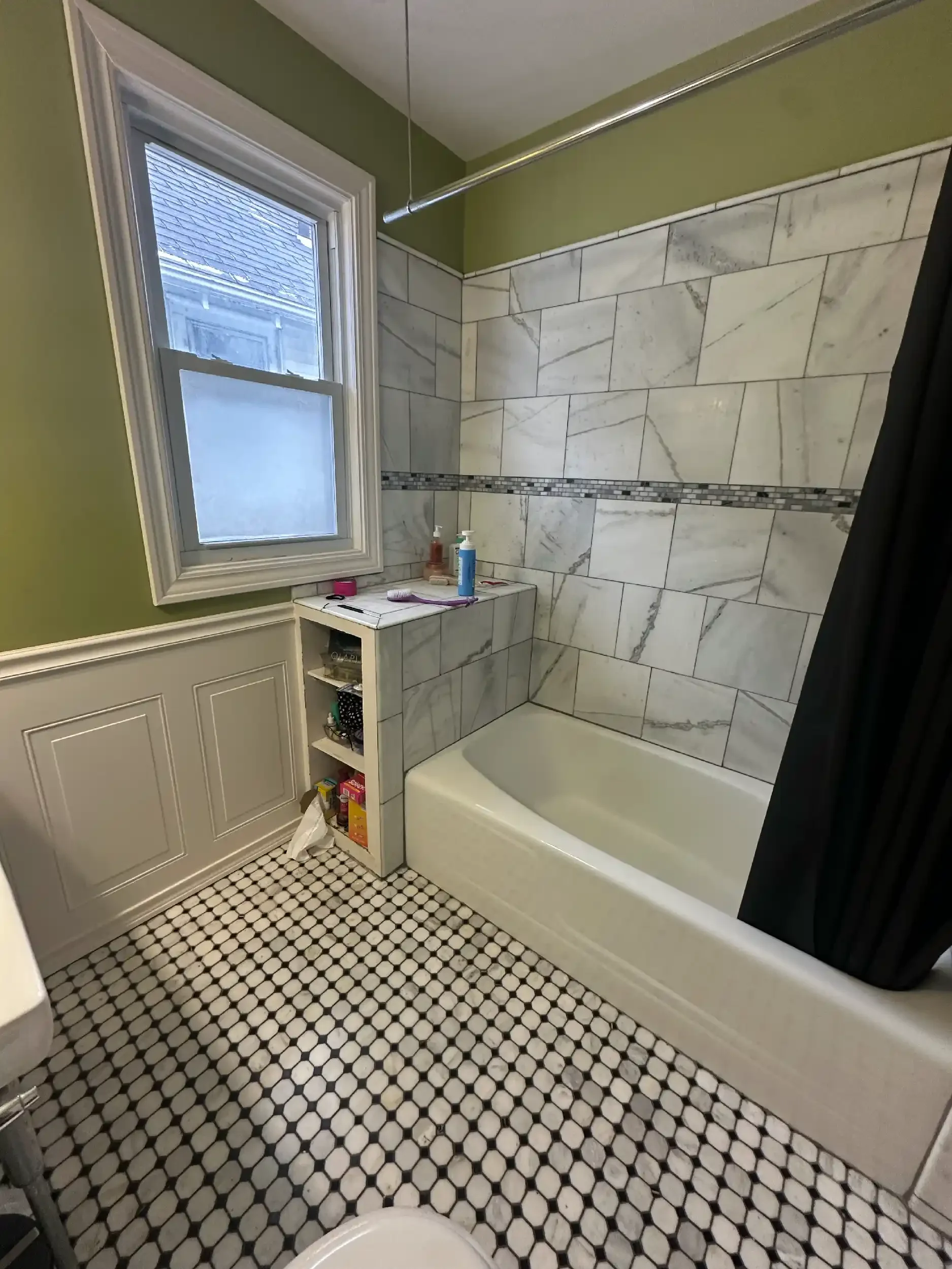 Bathroom with white hexagonal tile floor and marble tile shower wall. Green walls above the tile, with a window showing an exterior roof. A small white shelf under the window holds toiletries and other small items.
