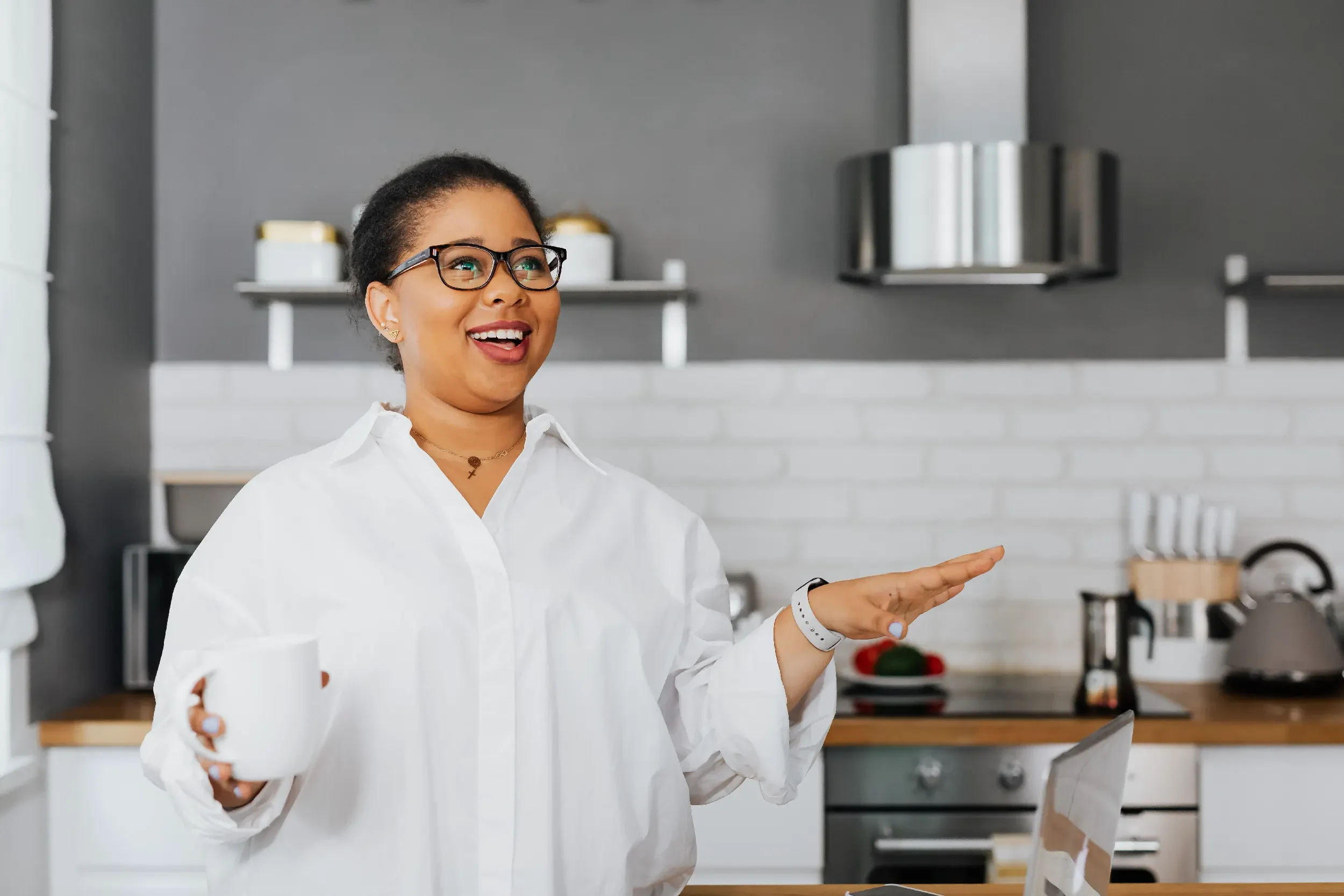 A woman with dark hair in a bun, wearing glasses and a white shirt, smiling and gesturing with her hand in a modern kitchen.